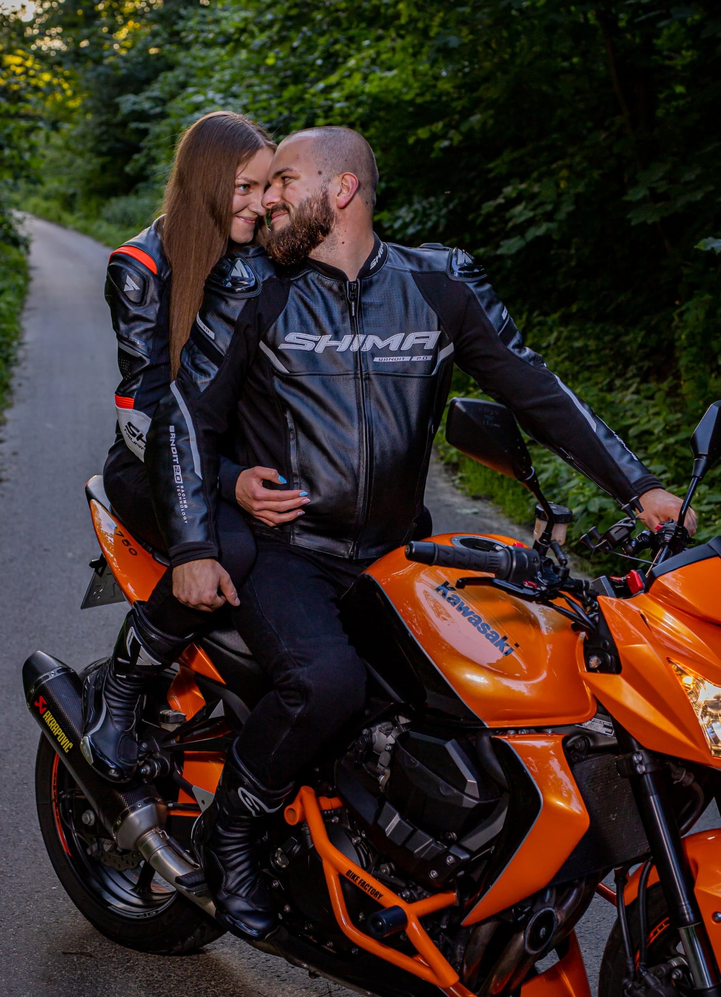 Para w skórzanych strojach motocyklowych na pomarańczowym Kawasaki Z750, sfotografowana na leśnej drodze w ciepłym oświetleniu.