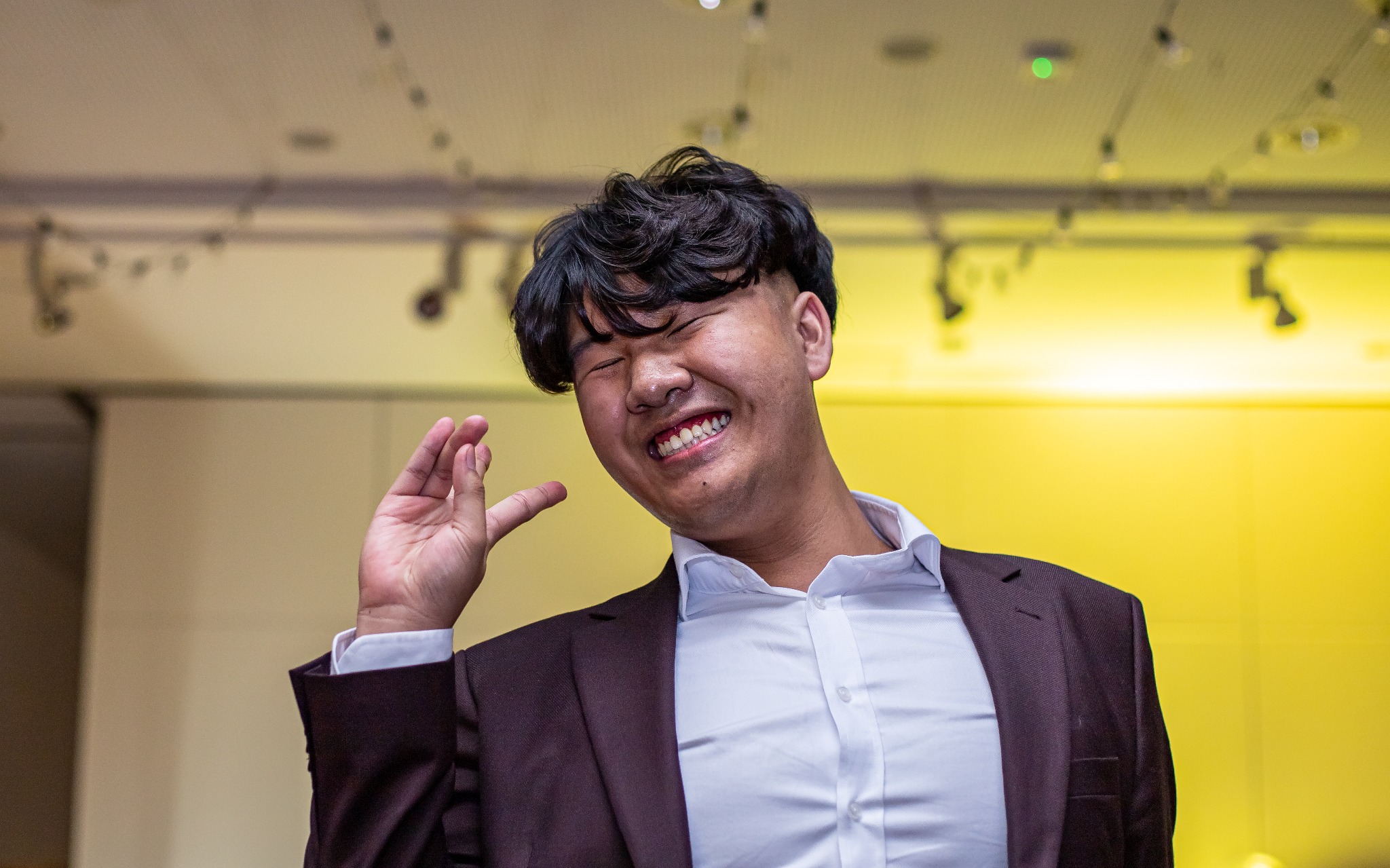 Uśmiechnięty mężczyzna w bordowej marynarce i białej koszuli, z zamkniętymi oczami, gestykuluje dłonią na tle żółtej ściany. Wyraźny uśmiech i ekspresja radości.