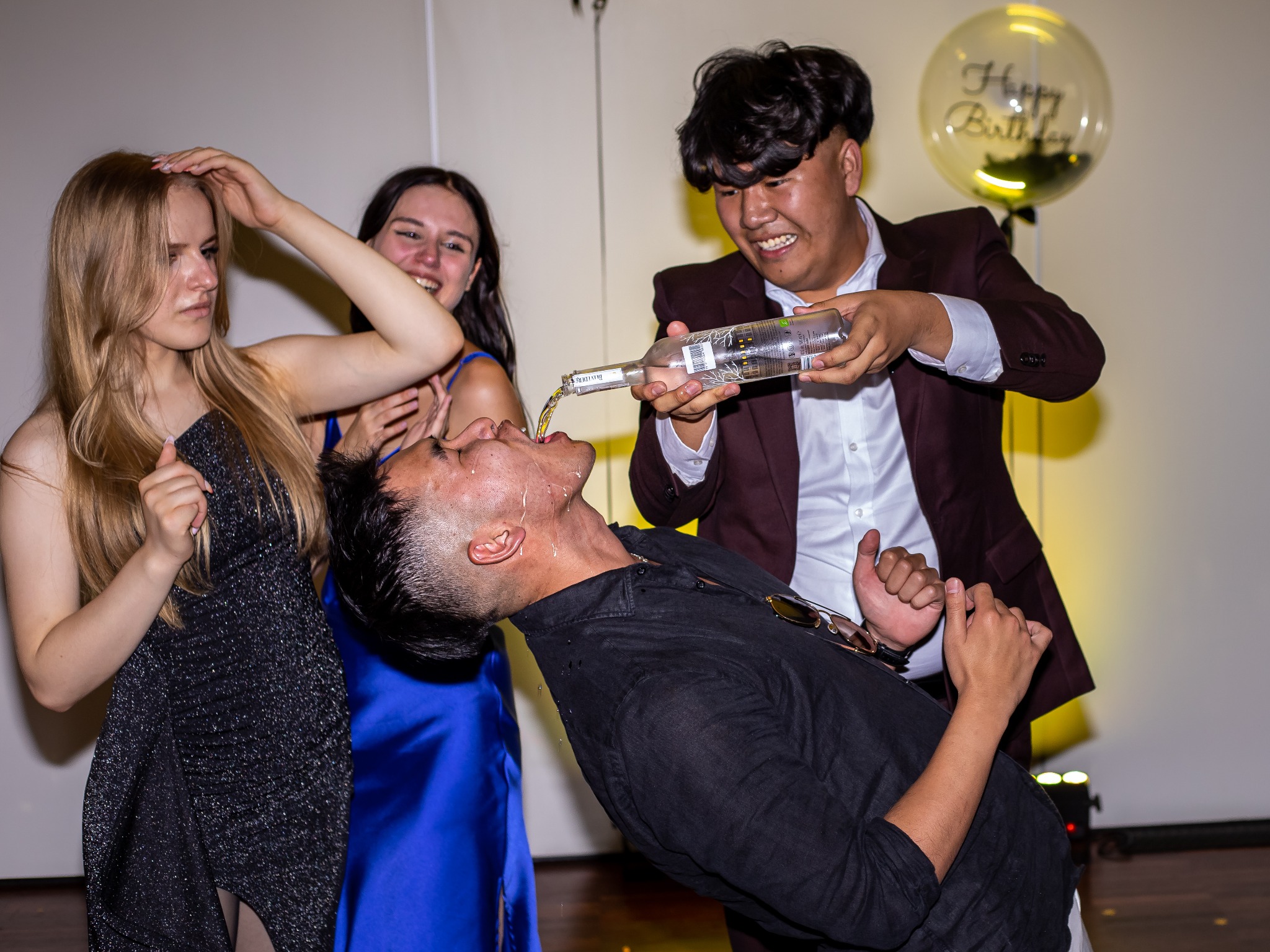 Mężczyzna pije alkohol wlewany mu do ust przez kolegę, podczas gdy dwie kobiety się śmieją. W tle balon z napisem 'Happy Birthday'.