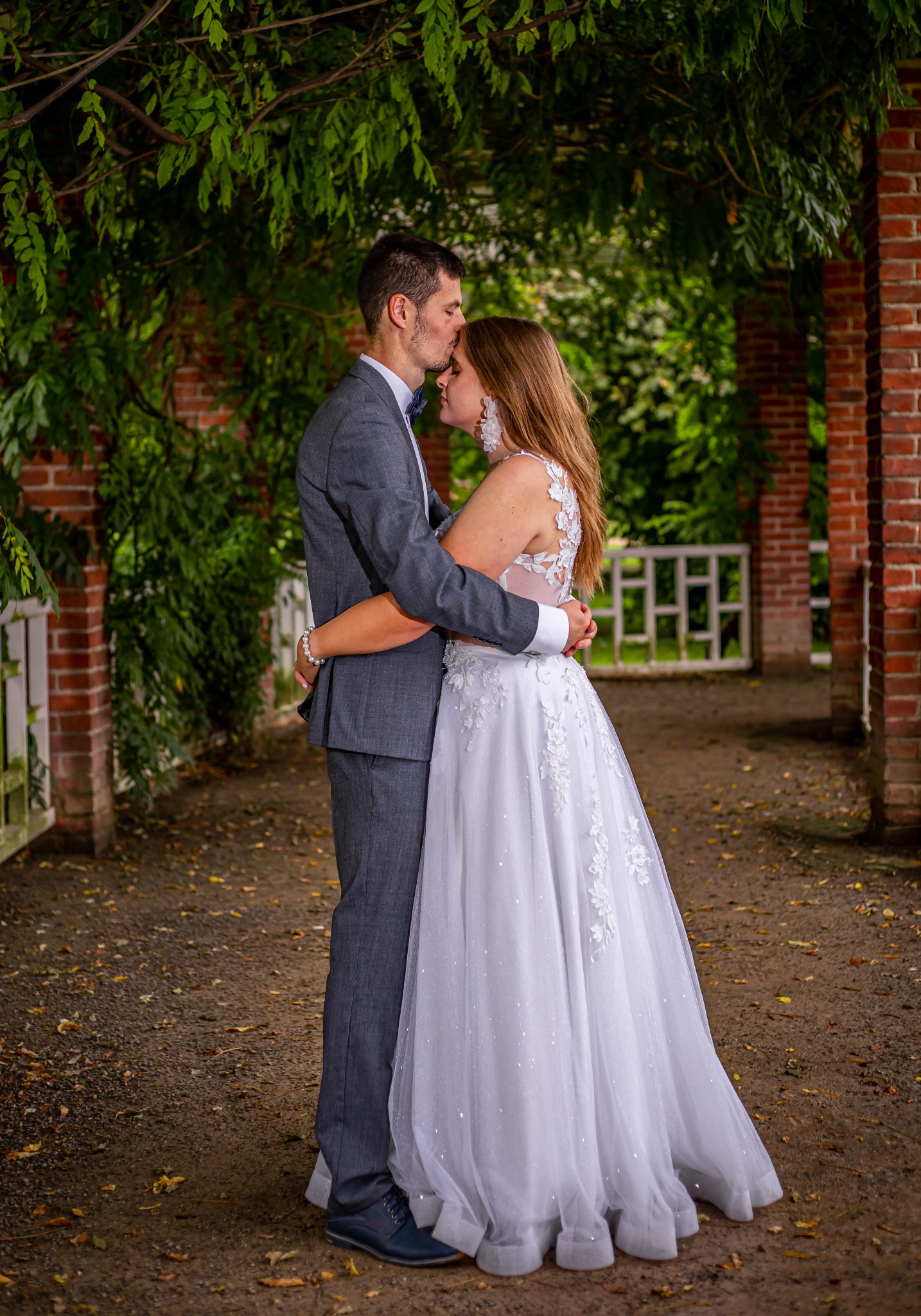 Para młoda w objęciach: on w szarym garniturze, ona w białej sukni z koronką, na tle ceglanej architektury i zieleni. Mężczyzna całuje kobietę w czoło. Ujęcie plenerowe.
