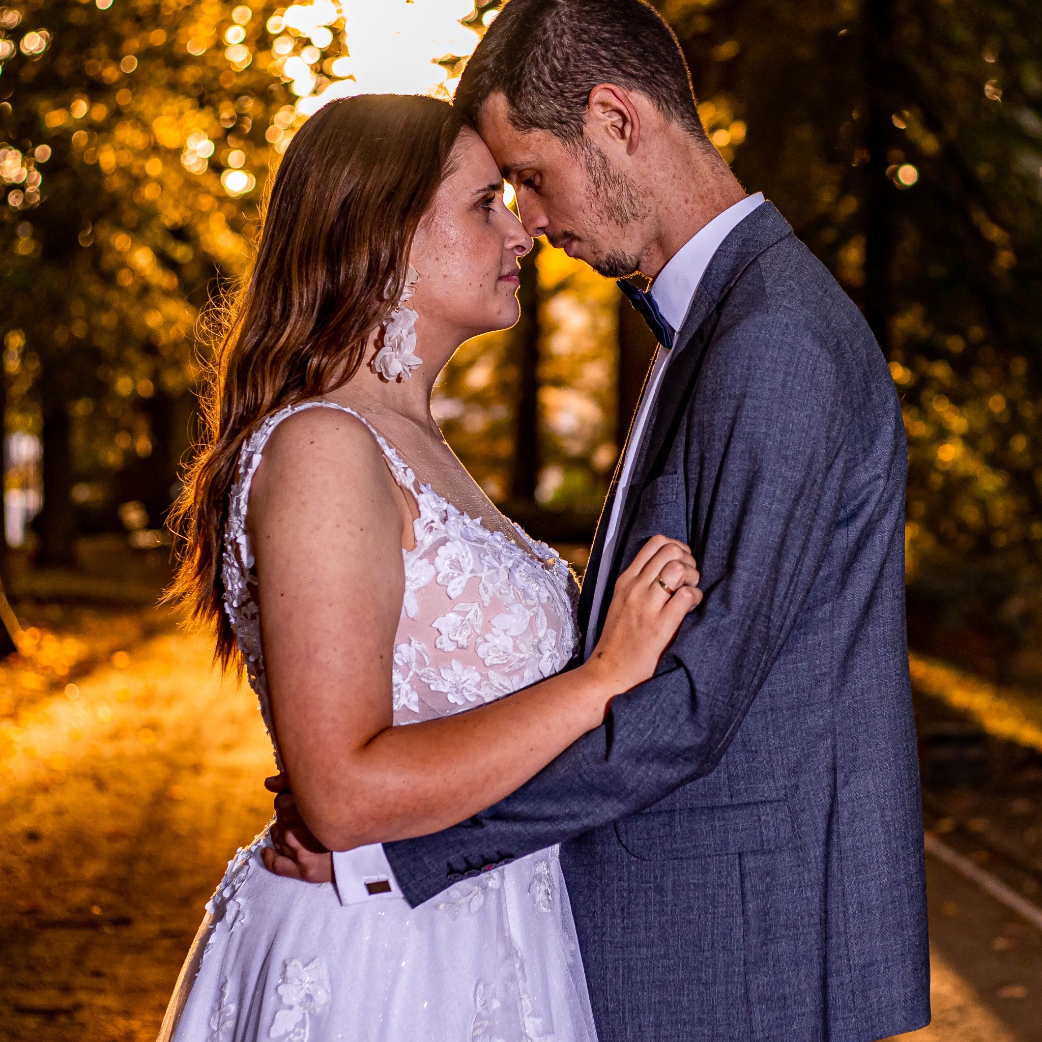 Zakochana para w objęciach na tle złotych świateł. Ona w białej sukni z koronką, on w szarym garniturze z muchą. Romantyczna atmosfera, bliskość i miłość.