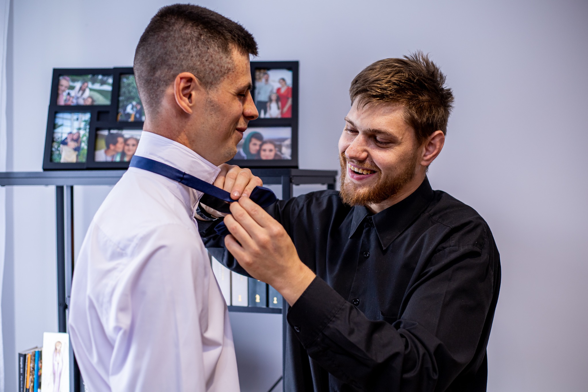 Uśmiechnięty mężczyzna w czarnej koszuli poprawia krawat drugiemu mężczyźnie w białej koszuli. W tle półka z ramkami ze zdjęciami.