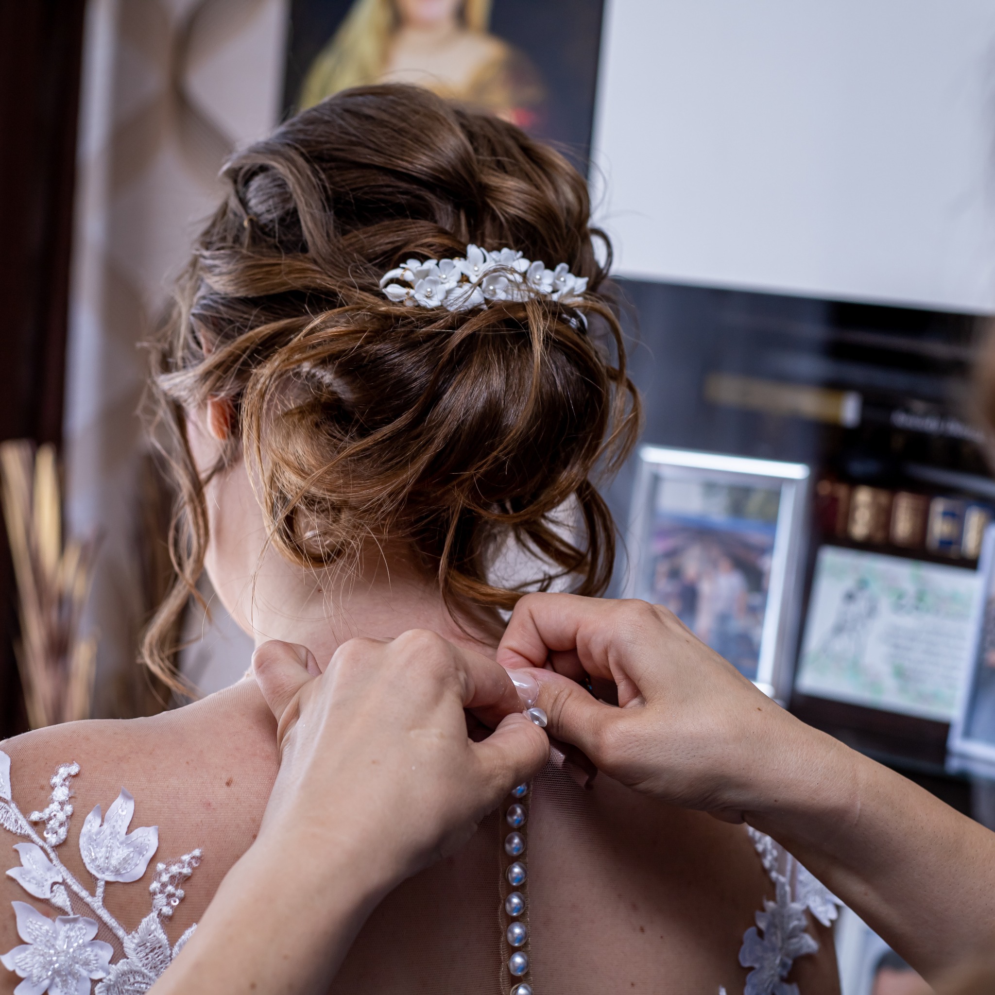Zapinanie sukni ślubnej z perłami na plecach panny młodej z upięciem włosów i ozdobą. Widoczne ręce pomagające zapiąć suknię. Tło rozmyte, z ramkami zdjęć.