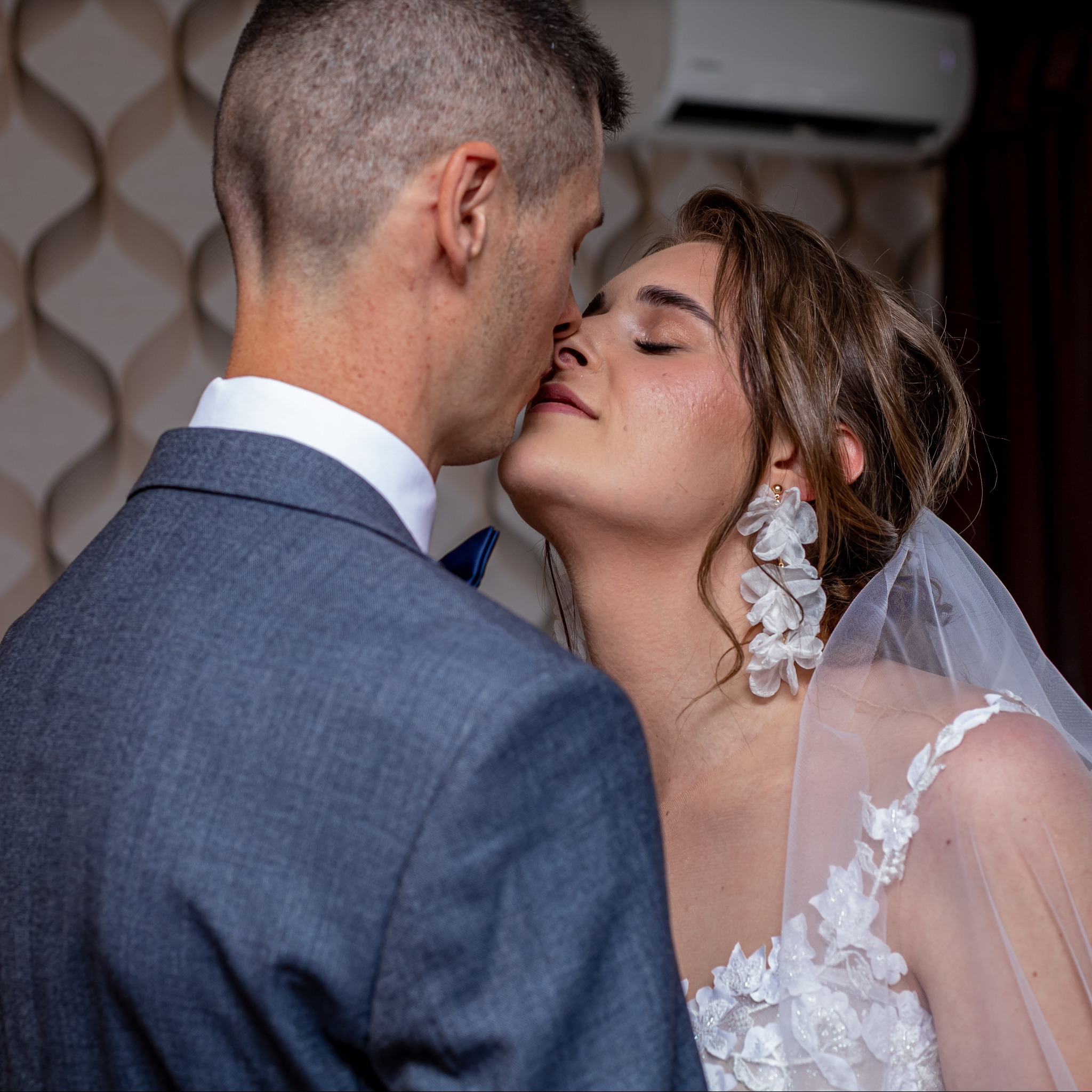 Zbliżenie na Pannę Młodą i Pana Młodego podczas pocałunku. Suknia z koronkowymi aplikacjami, welon i duże kolczyki w kształcie kwiatów. Elegancki garnitur.