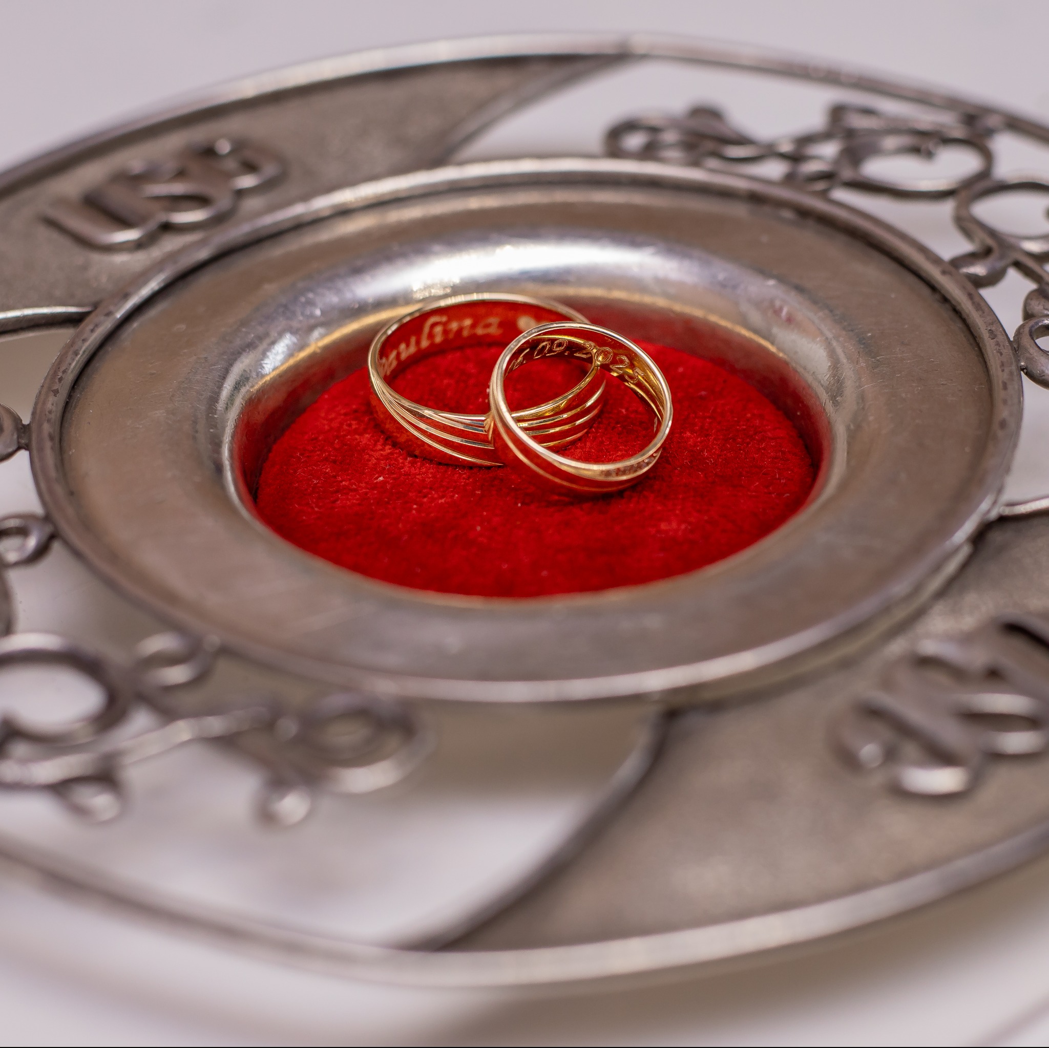 Dwa złote obrączki ślubne z wygrawerowanymi imionami i datą, leżące na czerwonej poduszce w srebrnej, zdobionej paterze. Makro, delikatne światło.