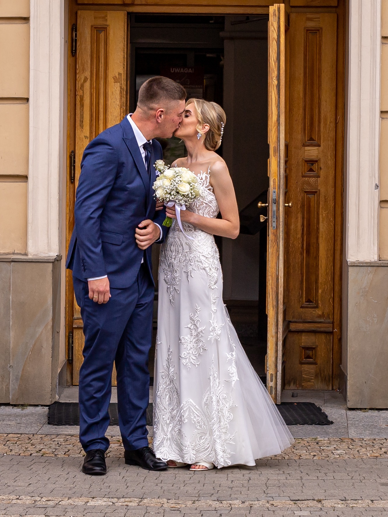 Pocałunek nowożeńców w drzwiach kościoła. Pan młody w granatowym garniturze, panna młoda w białej sukni z bukietem białych róż. Drewniane drzwi z tabliczką 'Uwaga!'.