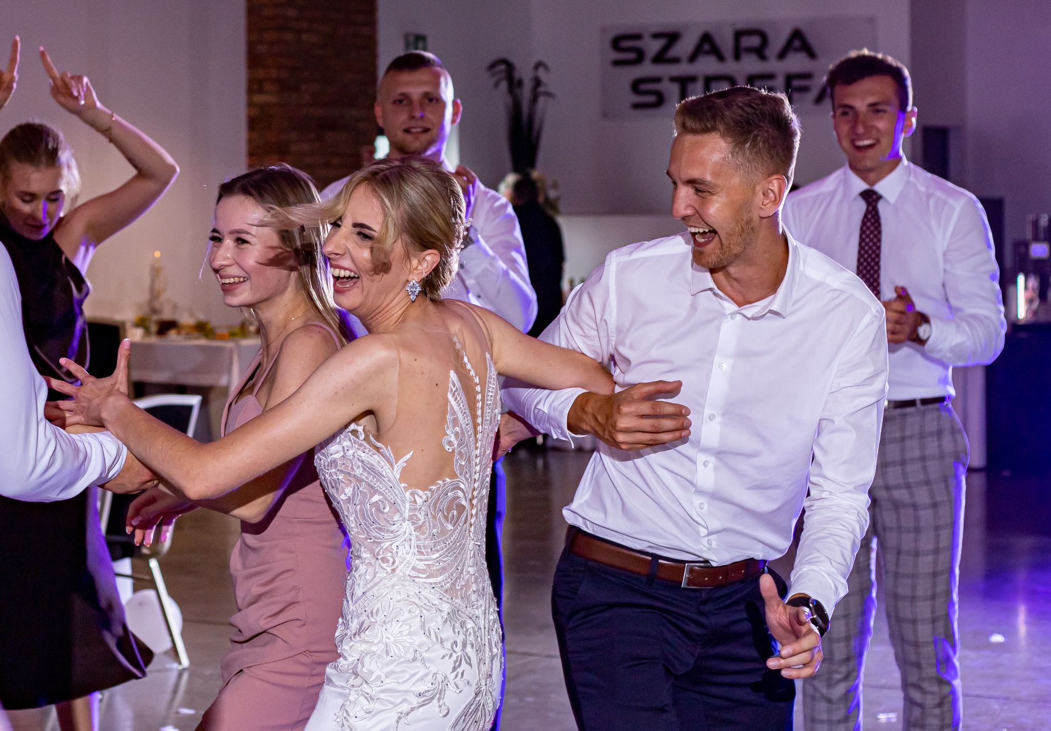 Rozbawiona para młoda w tańcu z gośćmi weselnymi. Panna młoda w koronkowej sukni, pan młody w białej koszuli. W tle uśmiechnięci goście i dekoracje sali.