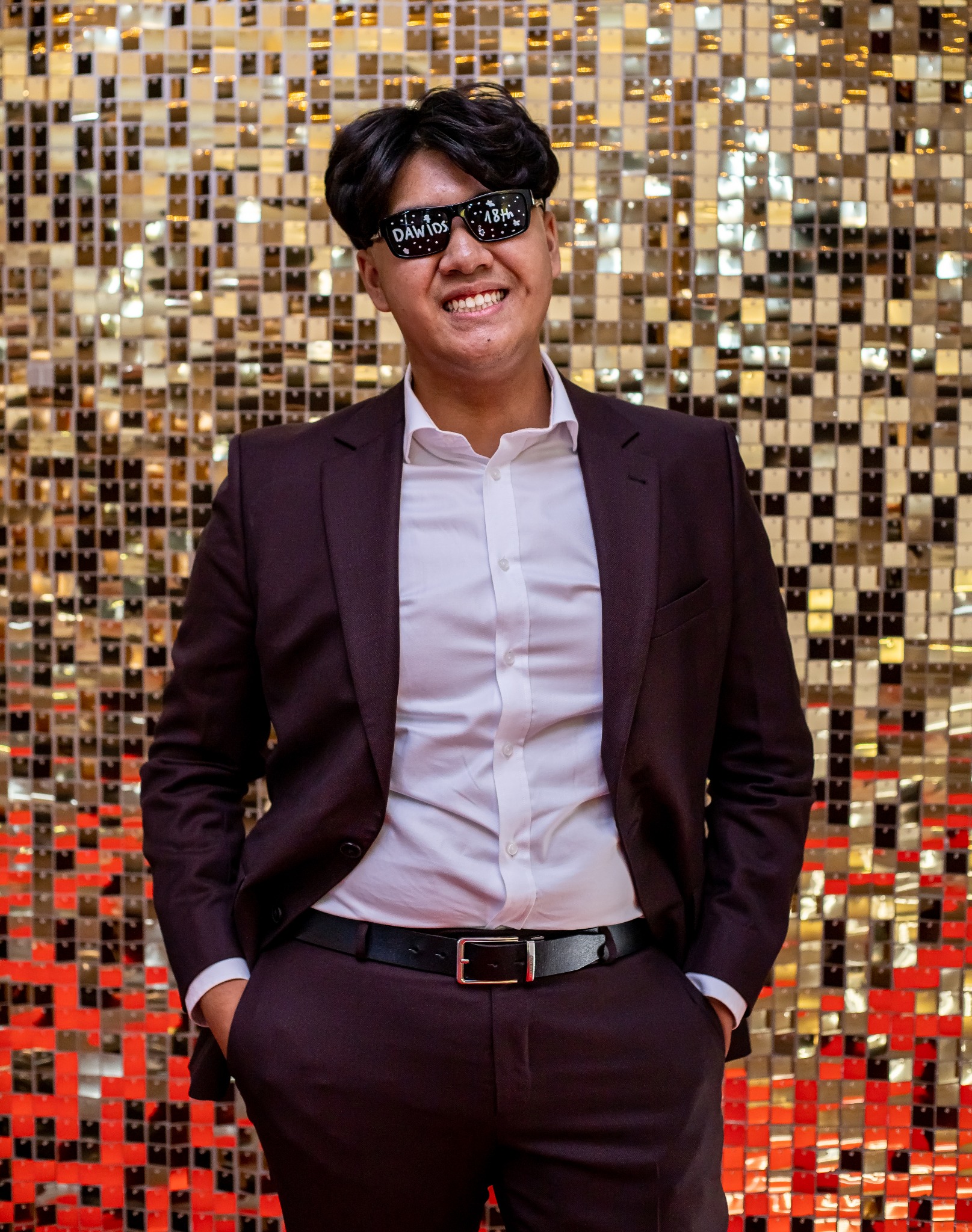 Mężczyzna w garniturze i okularach z napisem 'DAWIDS 18th' na tle błyszczącej ściany. Uśmiechnięty, z rękami w kieszeniach spodni.