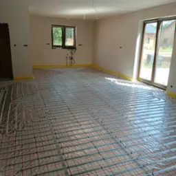 Białe rurki ogrzewania podłogowego ułożone na folii z czerwoną kratką w surowym pomieszczeniu z oknem i drzwiami balkonowymi.