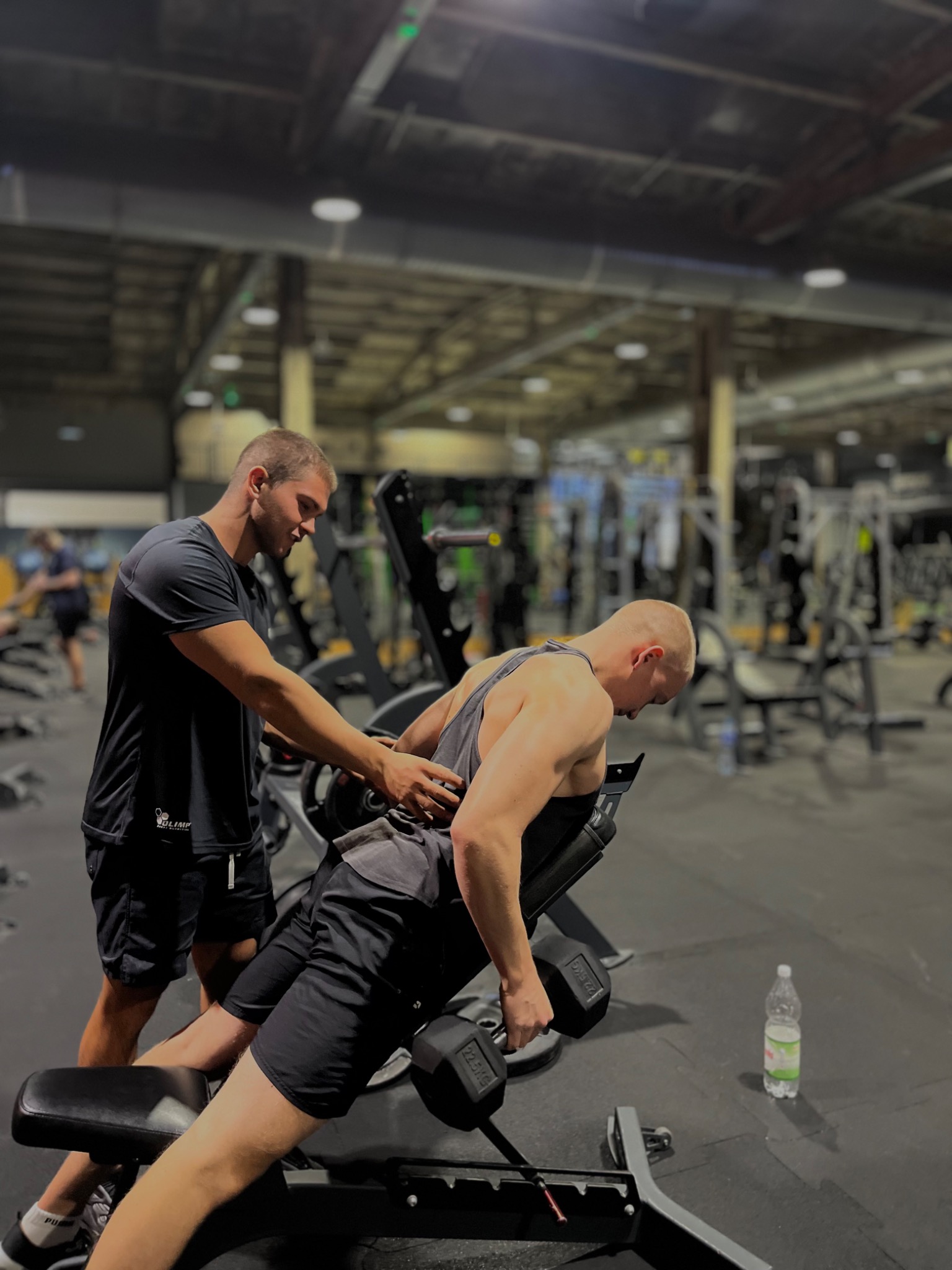 Mężczyzna z krótkimi włosami ćwiczy z hantlami na ławce skośnej w siłowni, a trener personalny go asekuruje.