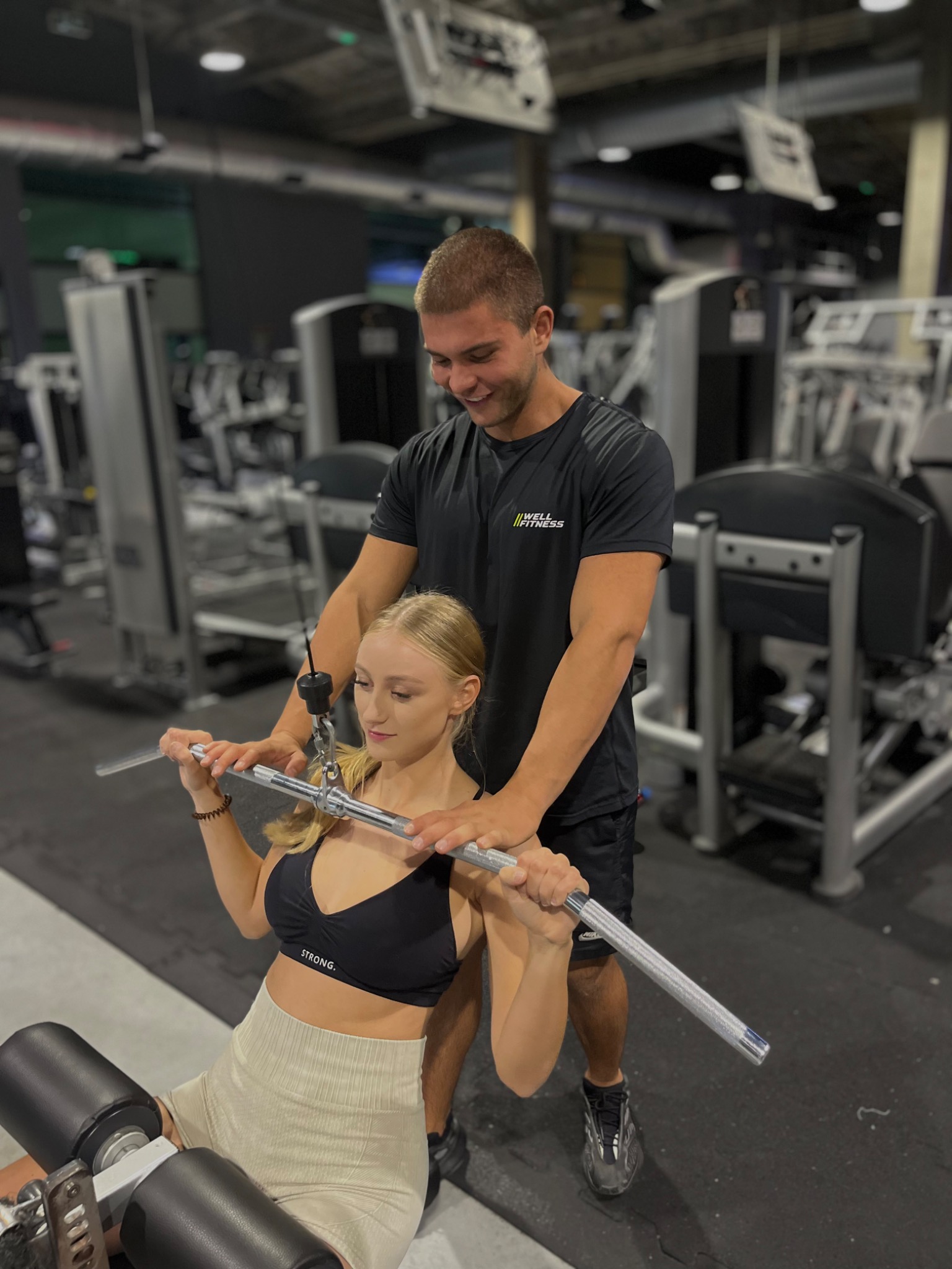 Kobieta ćwiczy na maszynie do wyciągania drążka pod nadzorem mężczyzny w siłowni, widoczne logo 'WELL FITNESS' na jego koszulce.