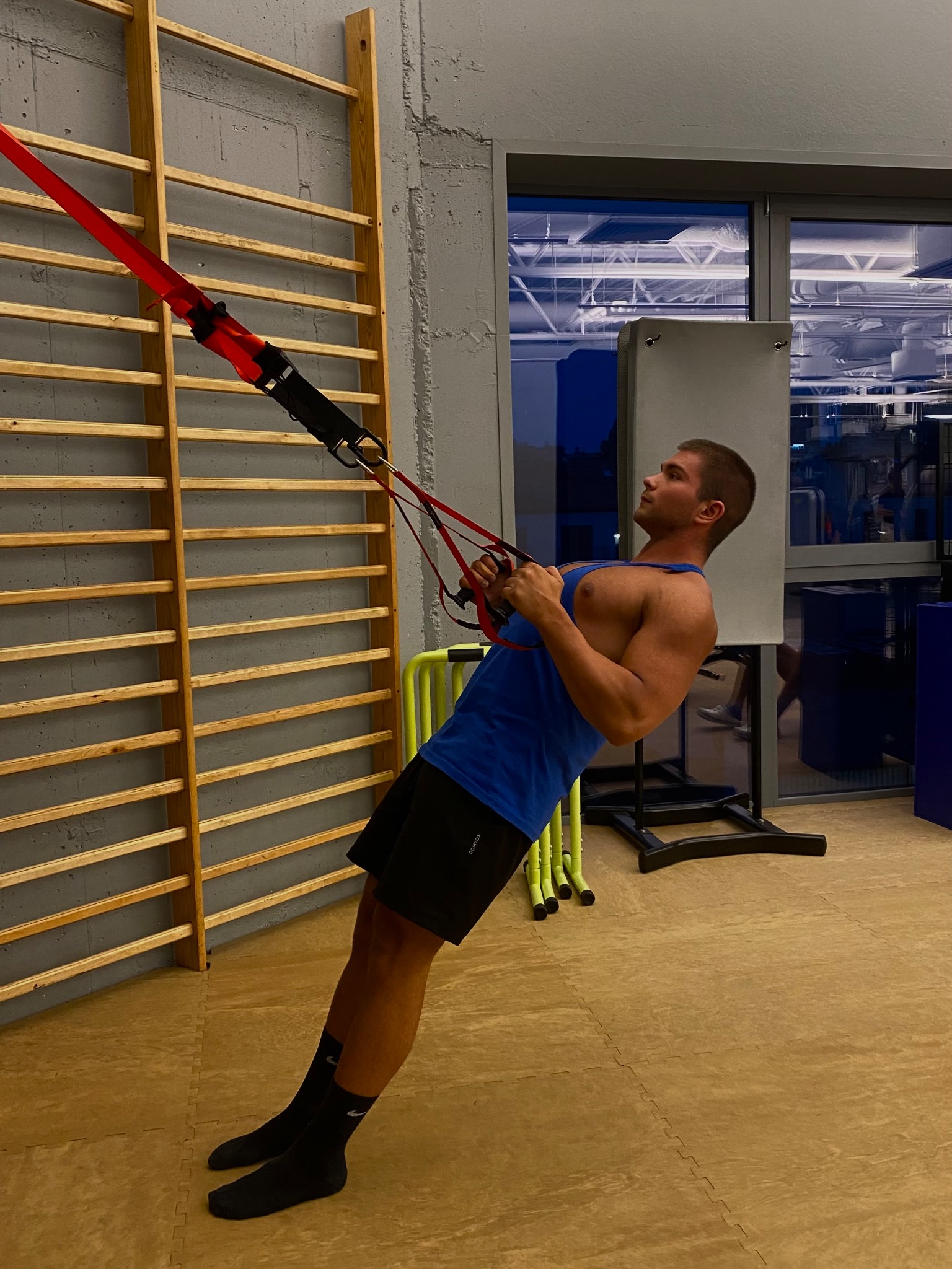 Mężczyzna w niebieskiej koszulce i czarnych spodenkach wykonuje ćwiczenie przy użyciu czerwonych taśm TRX zawieszonych na drabinkach gimnastycznych w siłowni.