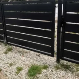 Nowoczesna, czarna brama wjazdowa z poziomymi szczeblami, zamontowana na posesji z kamienistym podłożem i fragmentami trawy.