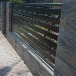 Nowoczesne ogrodzenie posesji z poziomych, metalowych profili w kolorze grafitowym, osadzone na słupkach i podmurówce z szarej, wzorzystej kostki.