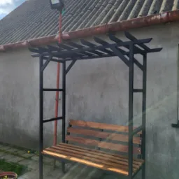 Metalowa konstrukcja z drewnianym siedziskiem ławki, czarne profile, brązowe deski, na zewnątrz, szara ściana w tle, oświetlenie zewnętrzne na dachu.