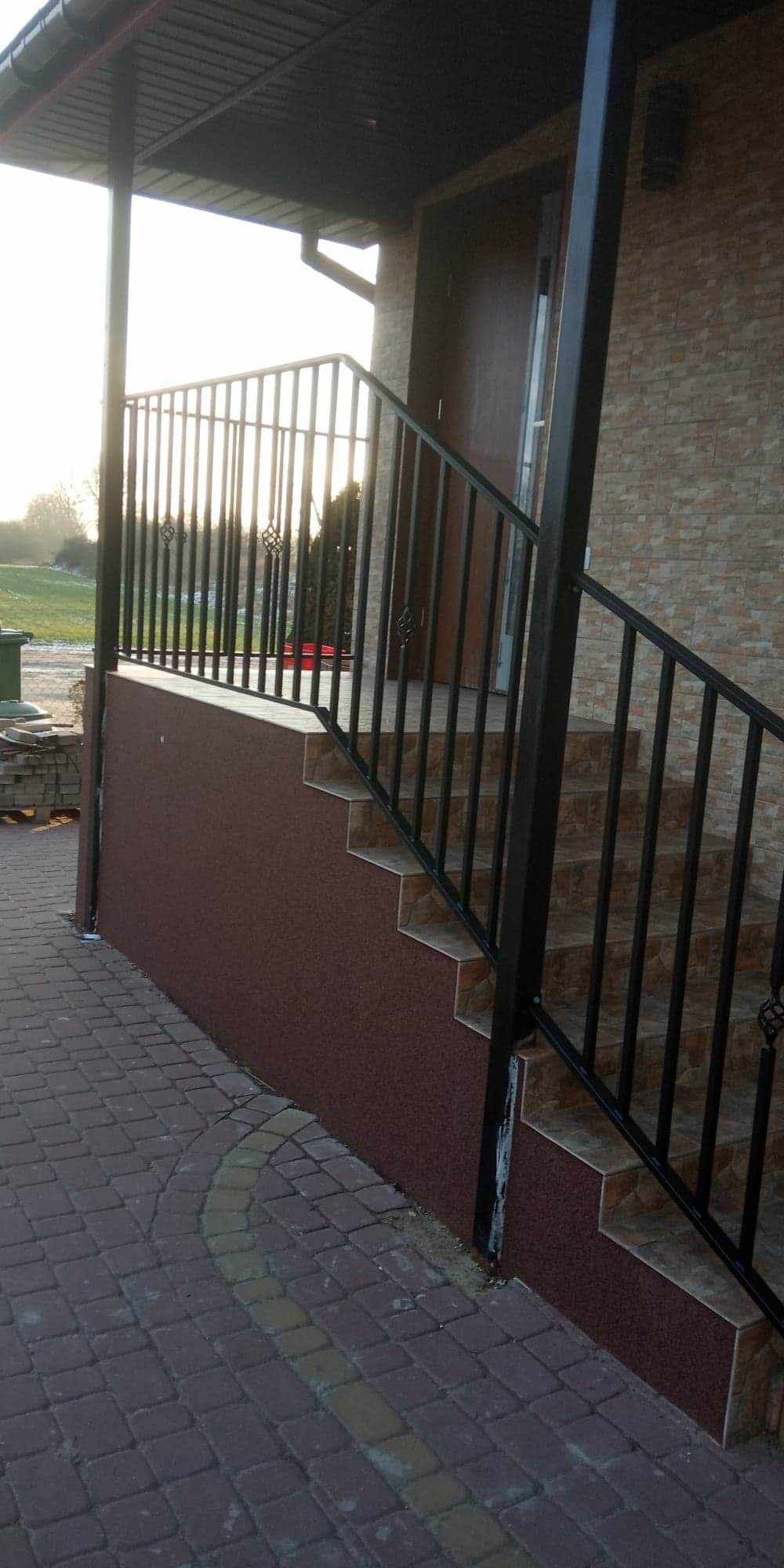 Zewnętrzne schody z brązowymi płytkami i czarną metalową balustradą z ozdobnymi elementami, prowadzące do drzwi wejściowych domu z elewacją z cegły.