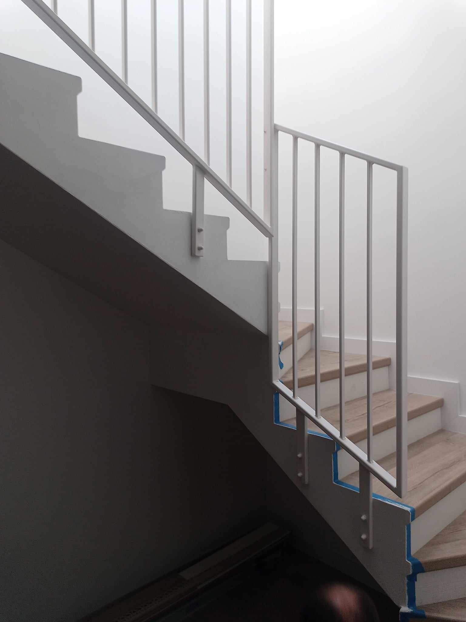 Białe schody z drewnianymi stopniami i metalową balustradą, zabezpieczone niebieską taśmą malarską podczas remontu.
