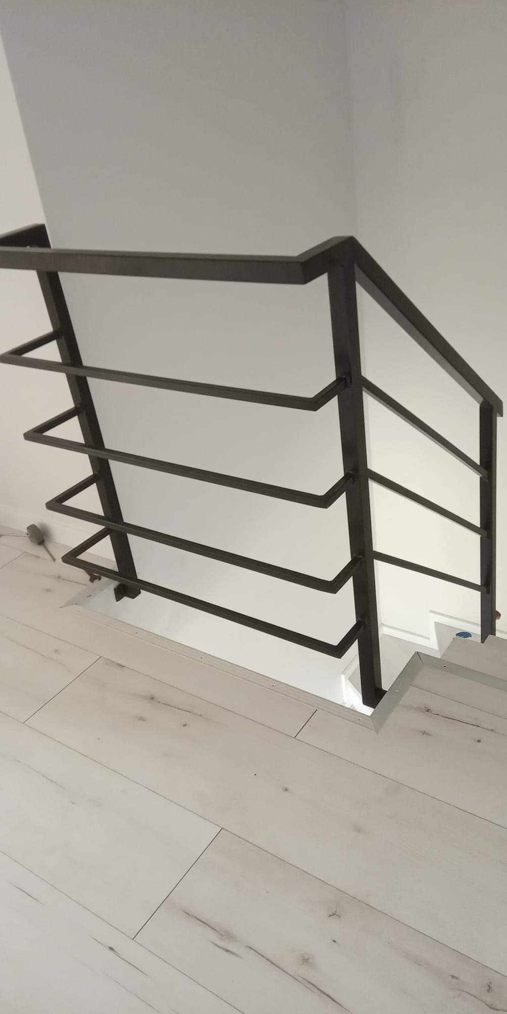 Nowoczesna, czarna balustrada schodowa z metalu, z poziomymi prętami o trójkątnym przekroju, zamontowana przy białej ścianie i schodach z jasnego drewna, widok z góry.