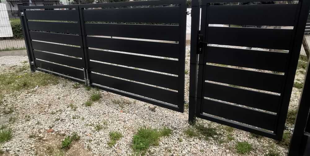 Nowoczesna, czarna brama wjazdowa z poziomymi szczeblami, zamontowana na posesji z kamienistym podłożem i fragmentami trawy.