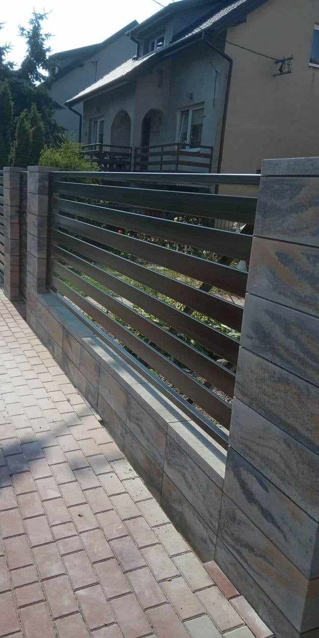 Nowoczesne ogrodzenie posesji z poziomych, metalowych profili w kolorze grafitowym, osadzone na słupkach i podmurówce z szarej, wzorzystej kostki.