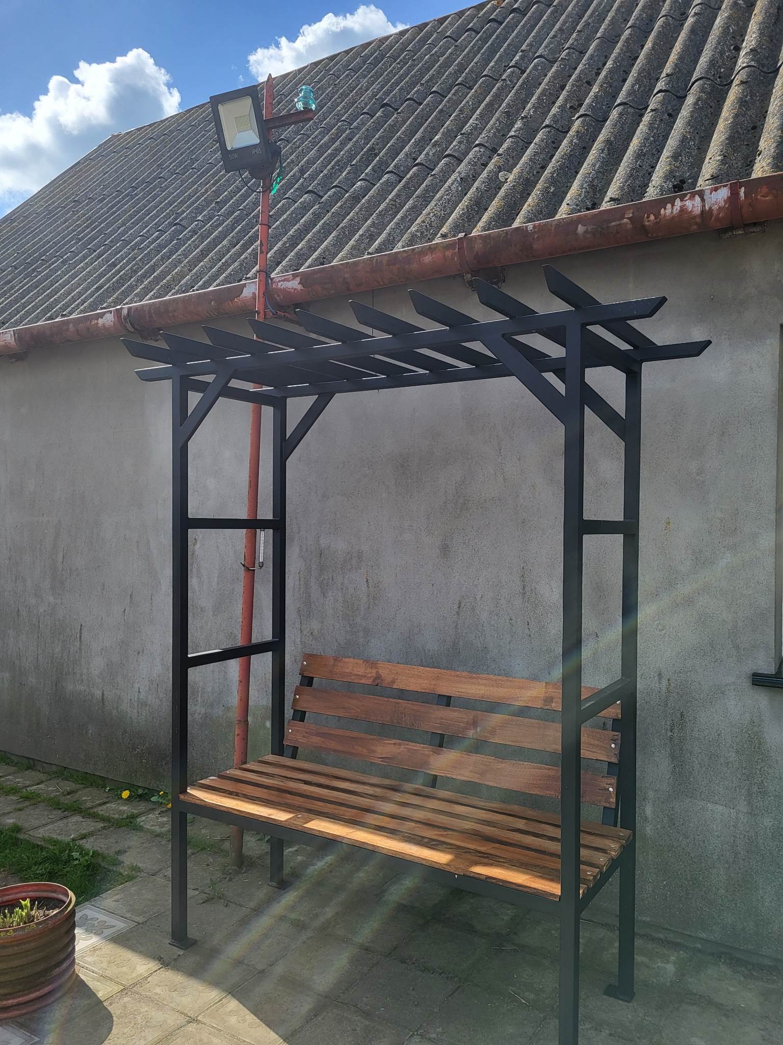 Metalowa konstrukcja z drewnianym siedziskiem ławki, czarne profile, brązowe deski, na zewnątrz, szara ściana w tle, oświetlenie zewnętrzne na dachu.