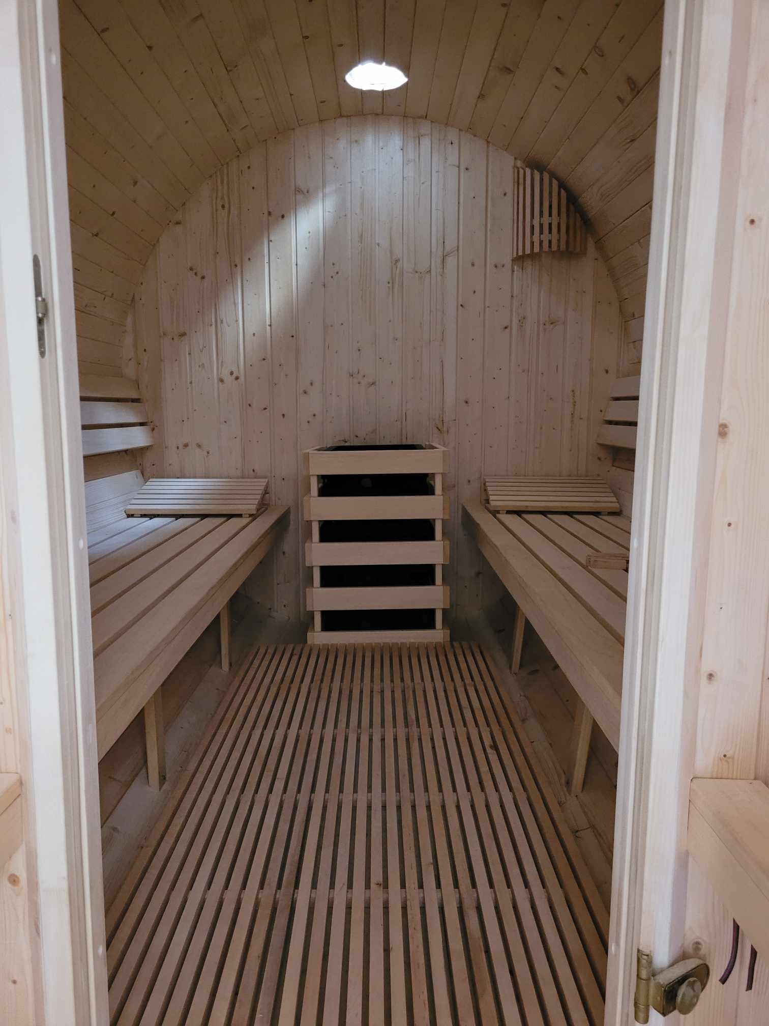Wnętrze drewnianej kabiny z piecem, ławkami i podłogą z listewek, oświetlone lampą sufitową, widok przez otwarte drzwi.