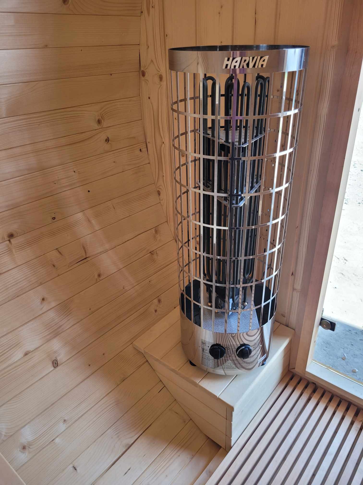 Wnętrze sauny z piecem elektrycznym Harvia w metalowej obudowie, drewniane ściany i podłoga z desek, widoczne regulatory pieca i wejście do sauny.