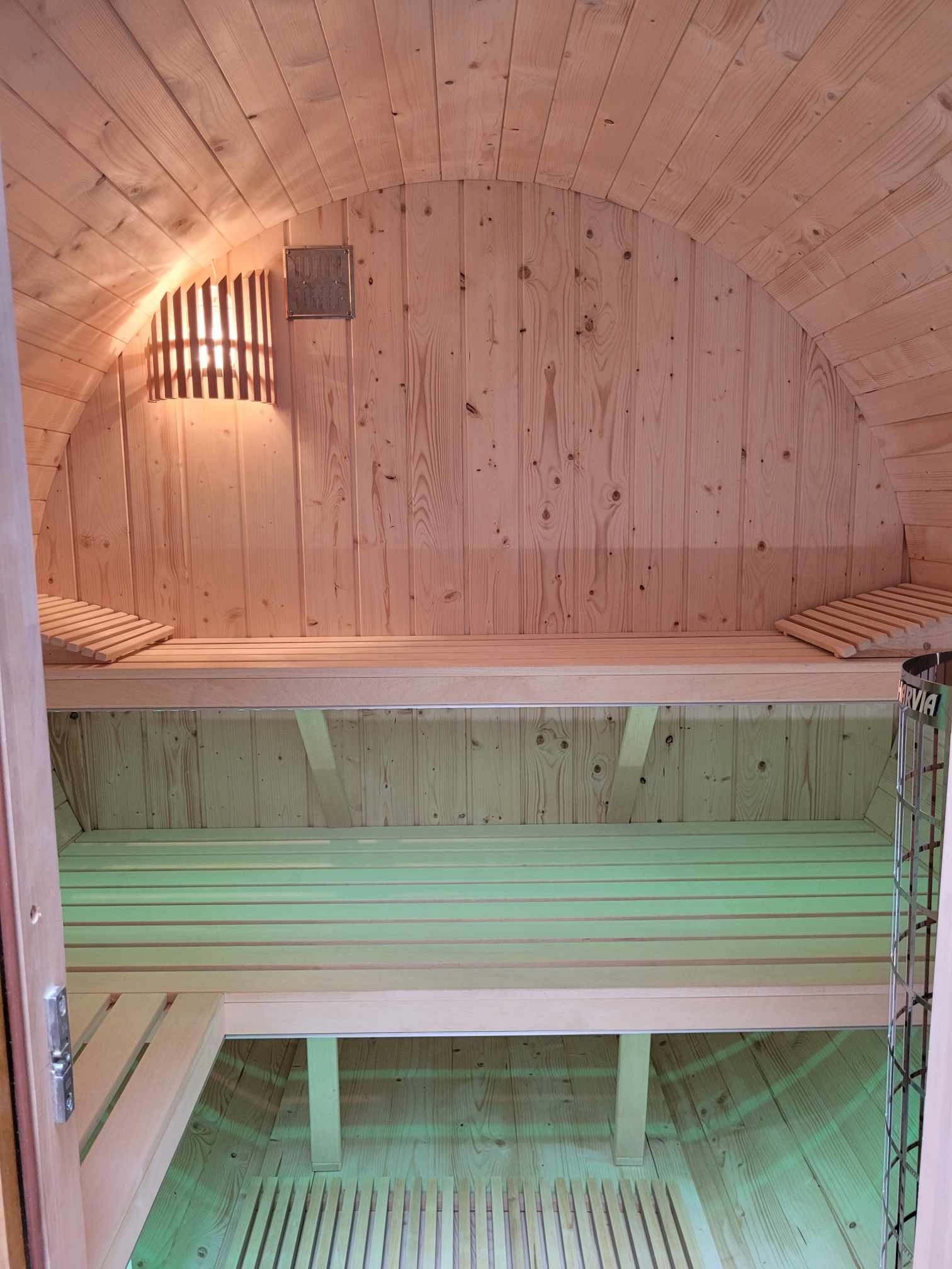 Wnętrze nowej, drewnianej sauny beczki z oświetleniem ściennym i metalowym piecem Harvia, widok na ławy z podgłówkami i podłogę z drewnianych listew.