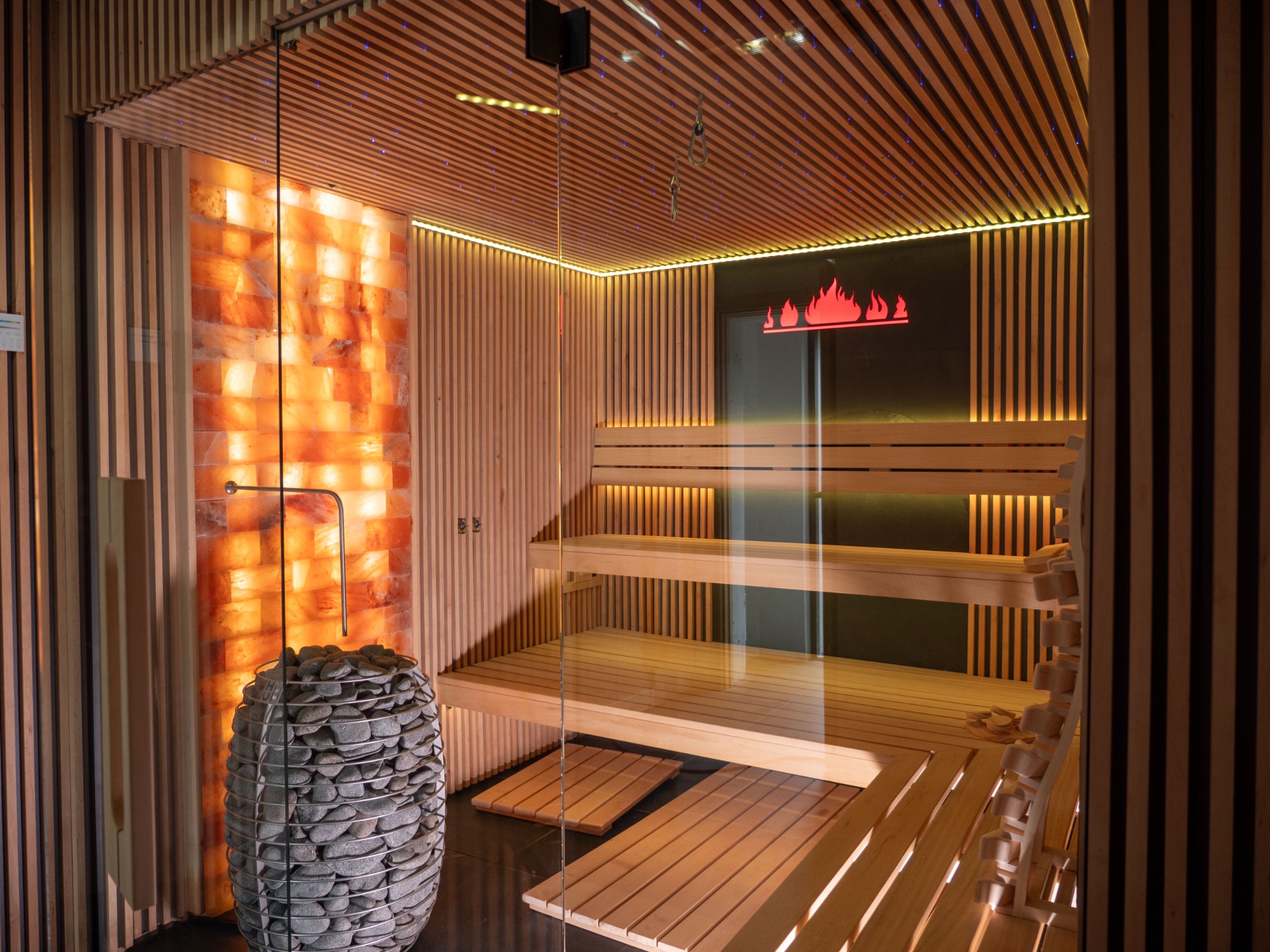 Wnętrze kabiny z drewnianymi ławkami, ścianą z bloków soli himalajskiej i piecem na kamienie w metalowej obudowie, podświetlane logo płomieni.