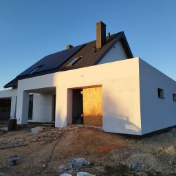 Witold Jablonski - Nowoczesny dom jednorodzinny w trakcie budowy, z białymi, gładkimi ścianami elewacyjnymi, ciemną dachówką i panelami słonecznymi, widoczne przygotowania do dalszych prac wykończeniowych na zewnątrz.