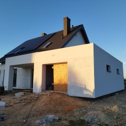 Witold Jablonski - Nowoczesny dom jednorodzinny z panelami słonecznymi na dachu, świeżo otynkowany, z prowizorycznym zamknięciem garażu z płyty OSB, pracownik budowlany z wiadrem farby na pierwszym planie.