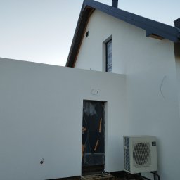 Witold Jablonski - Nowoczesny dom z białą elewacją w trakcie wykończenia, okno zabezpieczone folią, widoczna jednostka zewnętrzna klimatyzacji i drzwi wejściowe zakryte folią ochronną.