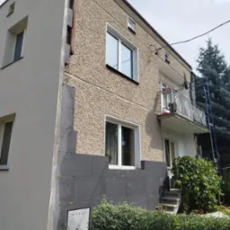 Dwukondygnacyjny budynek z fragmentem elewacji w trakcie prac: widoczna warstwa ocieplenia z szarego styropianu na narożniku, sąsiadująca z otynkowaną ścianą o fakturze baranka i świeżo pomalowaną...