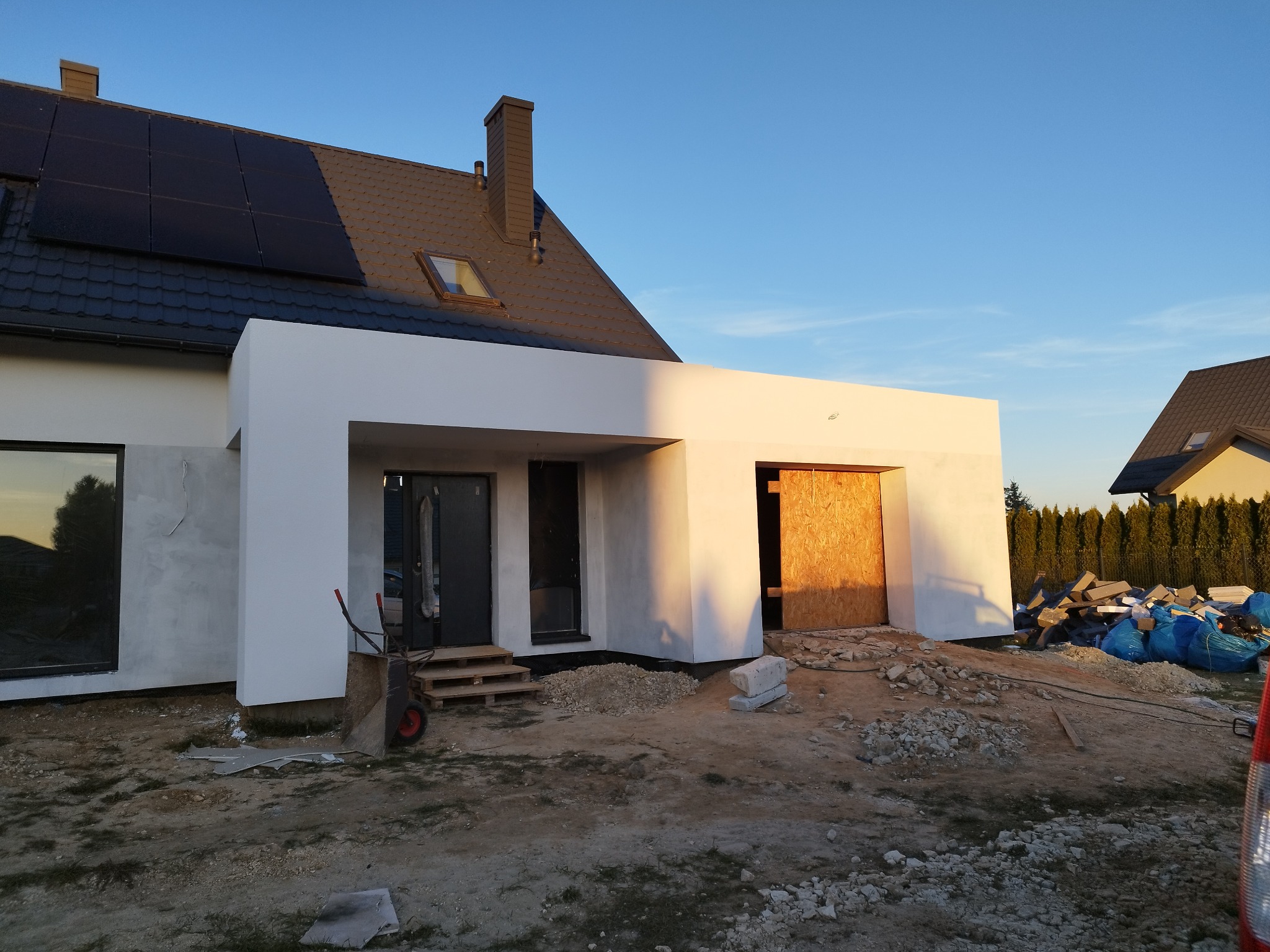 Nowoczesny dom jednorodzinny w trakcie prac elewacyjnych, z widocznymi panelami słonecznymi na dachu, świeżo otynkowanymi ścianami garażu i przygotowanym terenem wokół budynku.