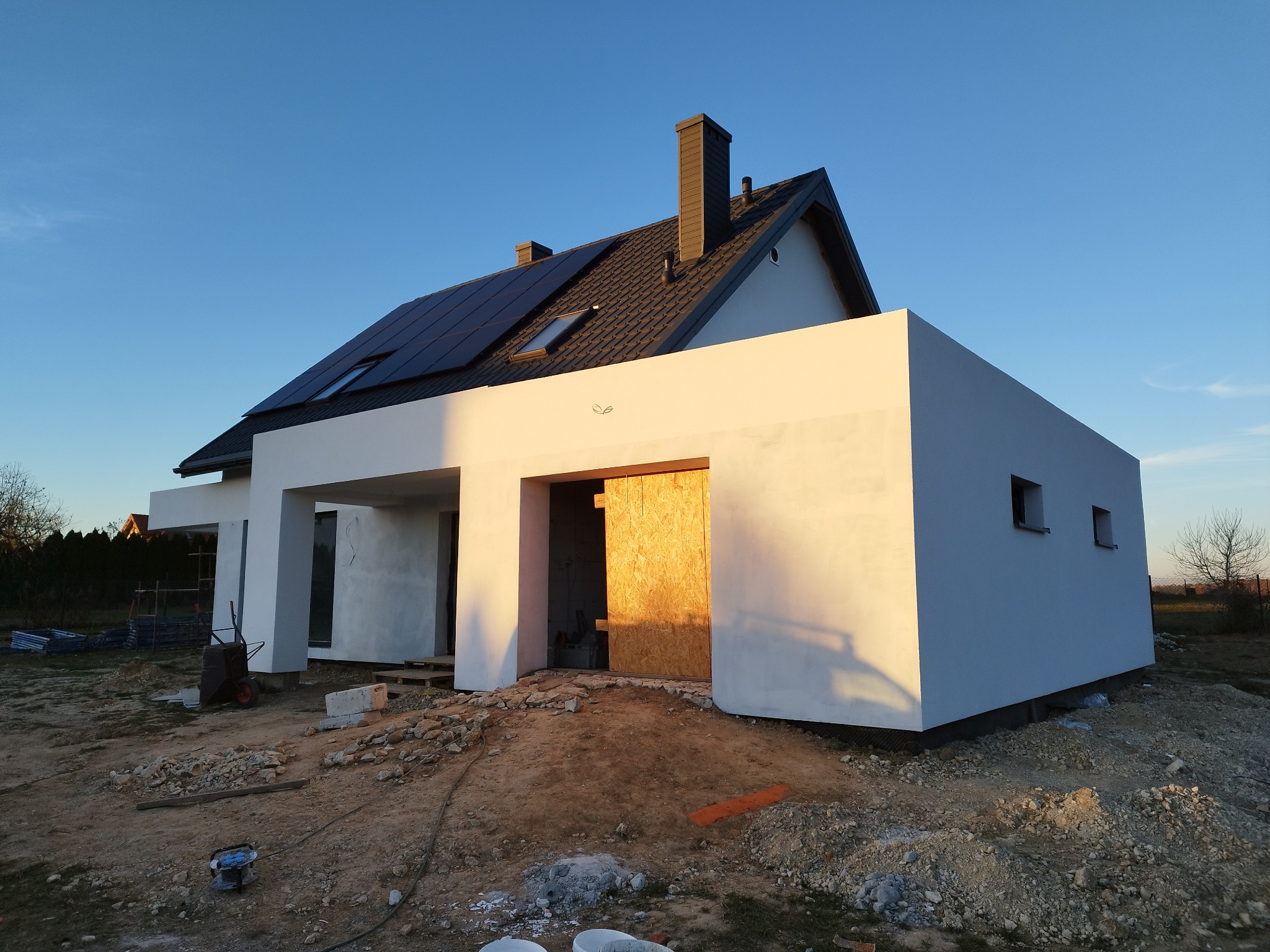 Nowoczesny dom jednorodzinny w trakcie budowy, z białymi, gładkimi ścianami elewacyjnymi, ciemną dachówką i panelami słonecznymi, widoczne przygotowania do dalszych prac wykończeniowych na zewnątrz.