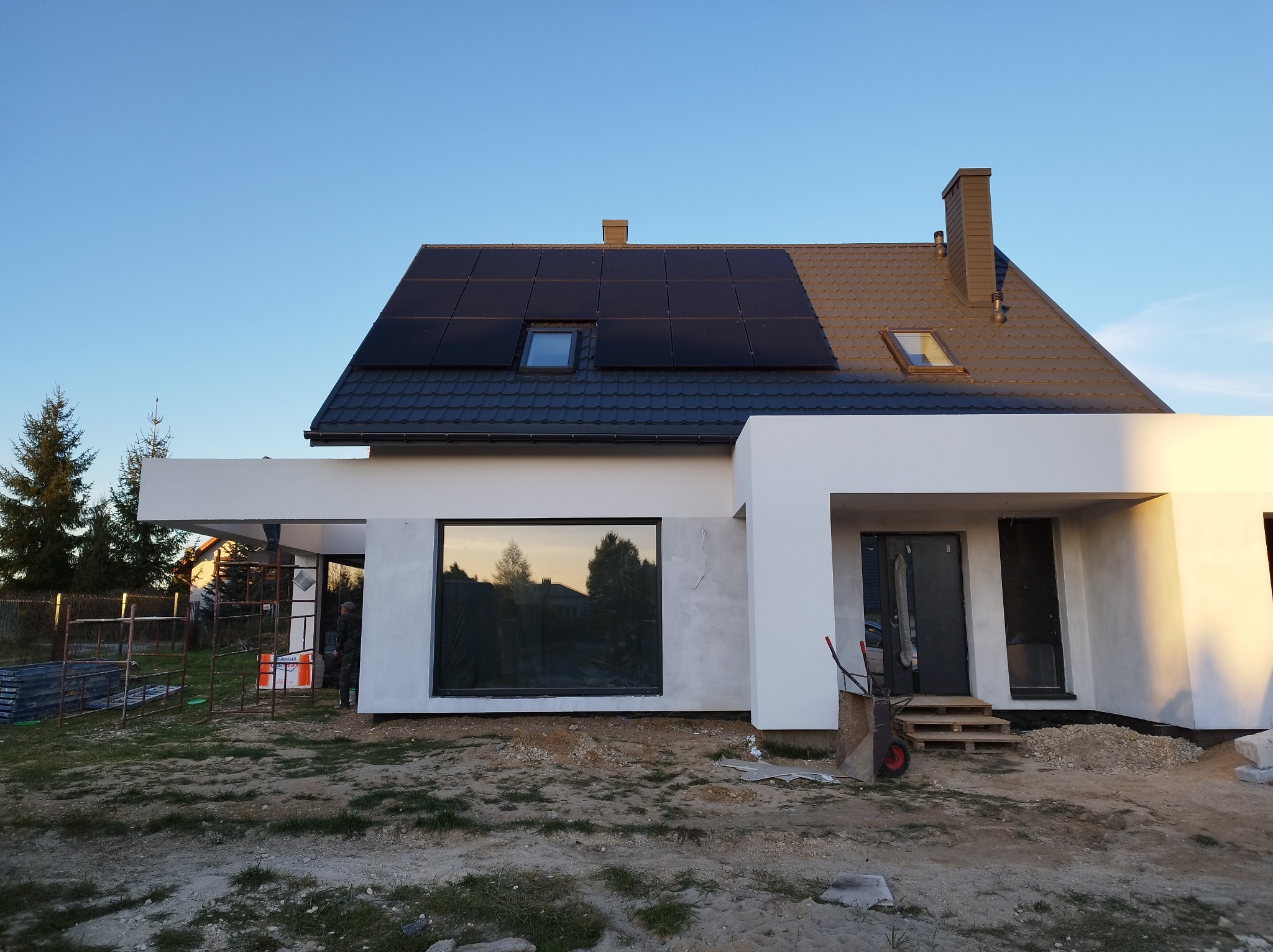 Nowoczesny dom jednorodzinny z panelami fotowoltaicznymi na dachu, elewacja w trakcie prac wykończeniowych, widoczne rusztowania i materiały budowlane.