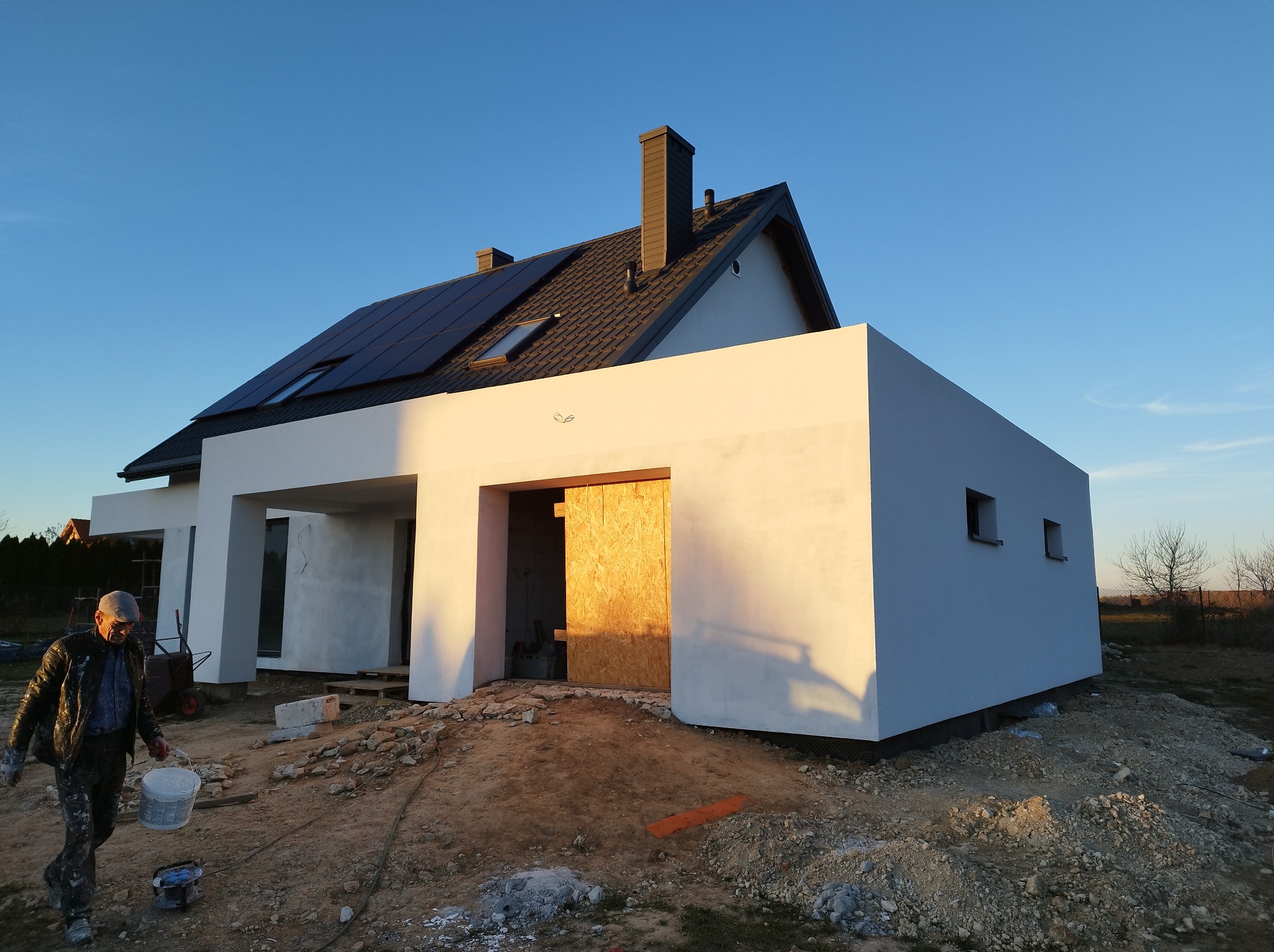 Nowoczesny dom jednorodzinny z panelami słonecznymi na dachu, świeżo otynkowany, z prowizorycznym zamknięciem garażu z płyty OSB, pracownik budowlany z wiadrem farby na pierwszym planie.