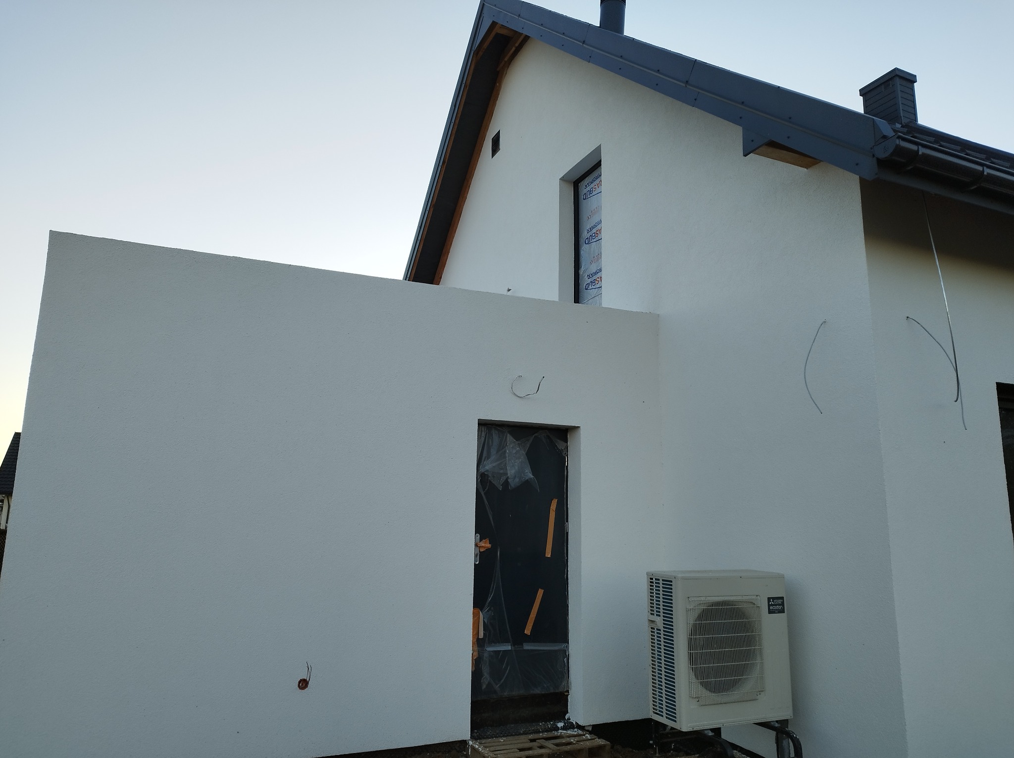 Nowoczesny dom z białą elewacją w trakcie wykończenia, okno zabezpieczone folią, widoczna jednostka zewnętrzna klimatyzacji i drzwi wejściowe zakryte folią ochronną.