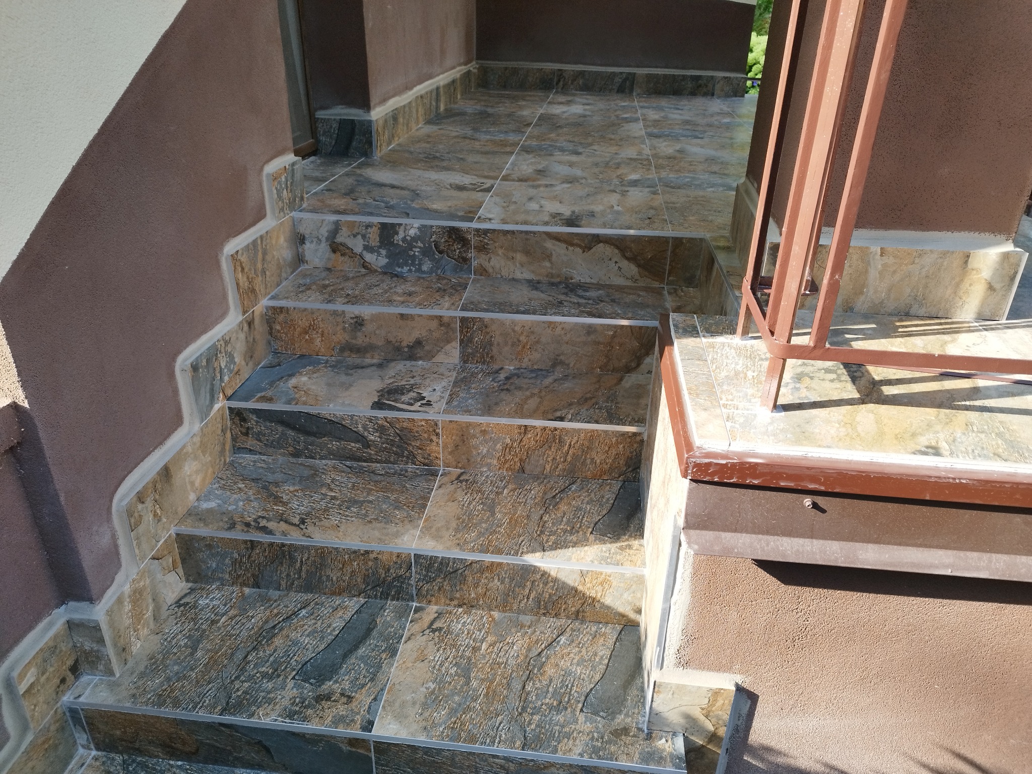 Schody wejściowe wyłożone płytkami imitującymi kamień, z widocznymi brązowymi ścianami o fakturze baranka i metalową balustradą.