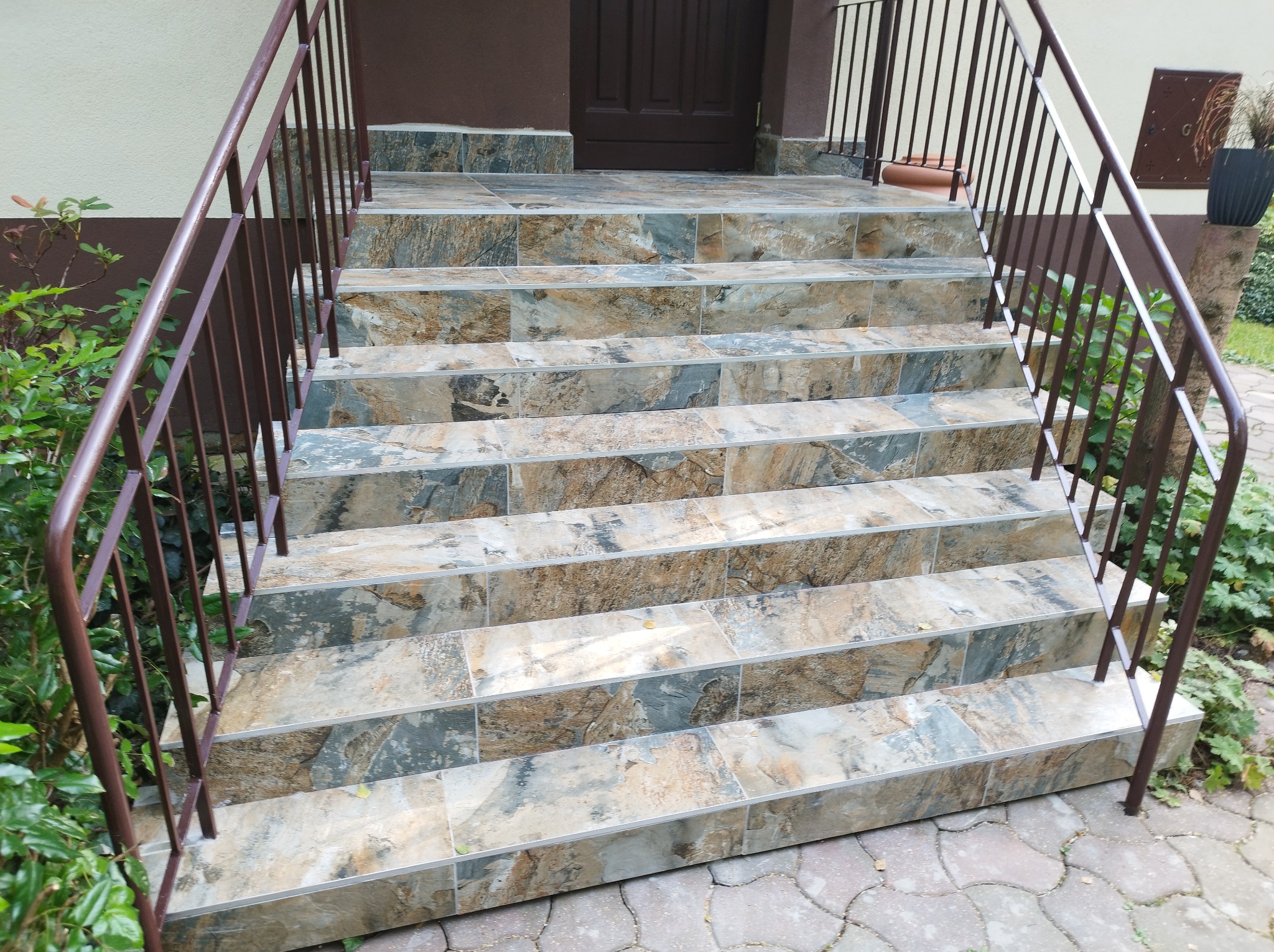 Wykończone schody wejściowe do domu z brązową balustradą i płytkami imitującymi kamień, prowadzące do ciemnobrązowych drzwi.