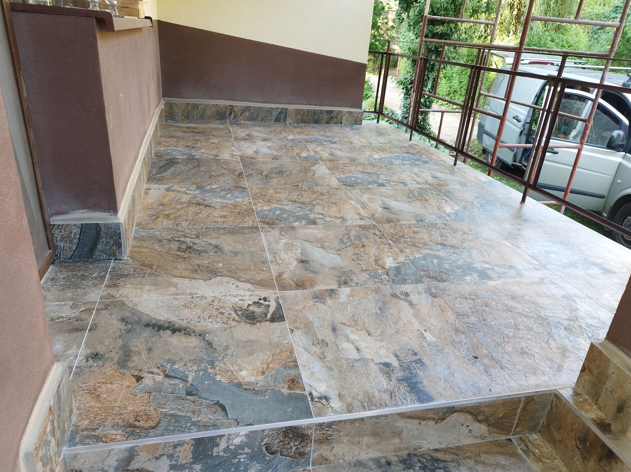 Wykończony taras z płytkami imitującymi kamień, brązowe ściany i balustrada z widocznym samochodem w tle.