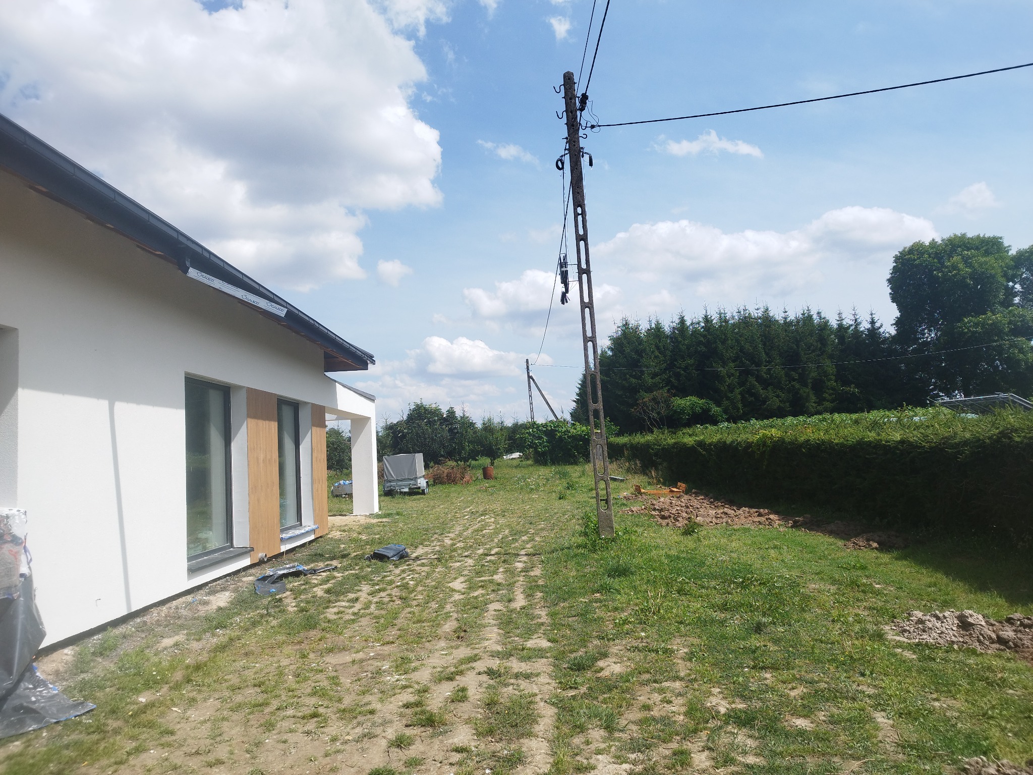 Nowoczesny dom jednorodzinny z białą elewacją i drewnianymi panelami okiennymi w trakcie wykończenia, widoczne zabezpieczenia na trawniku, słup energetyczny i przyczepa w tle.