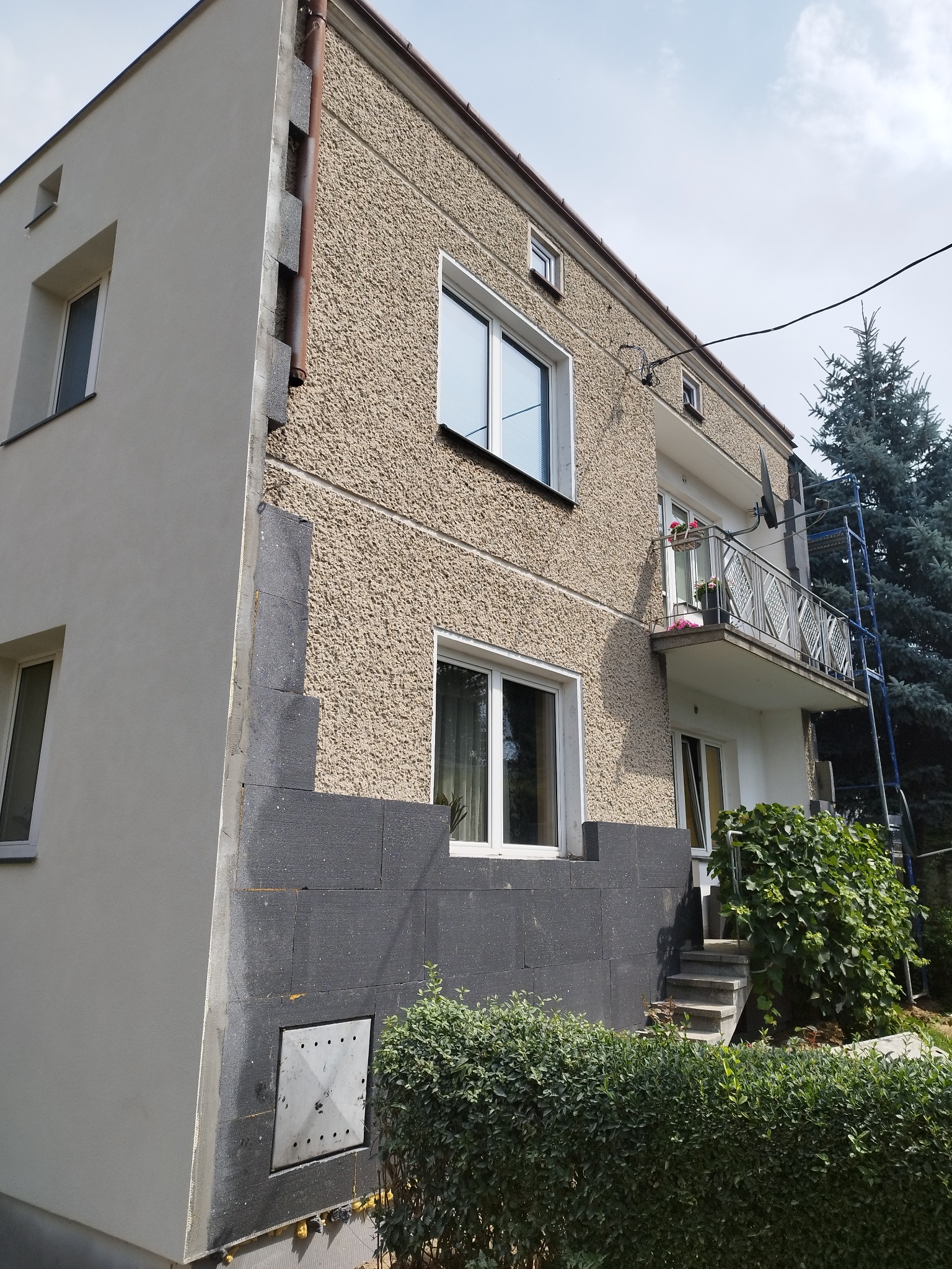 Dwukondygnacyjny budynek z fragmentem elewacji w trakcie prac: widoczna warstwa ocieplenia z szarego styropianu na narożniku, sąsiadująca z otynkowaną ścianą o fakturze baranka i świeżo pomalowaną...