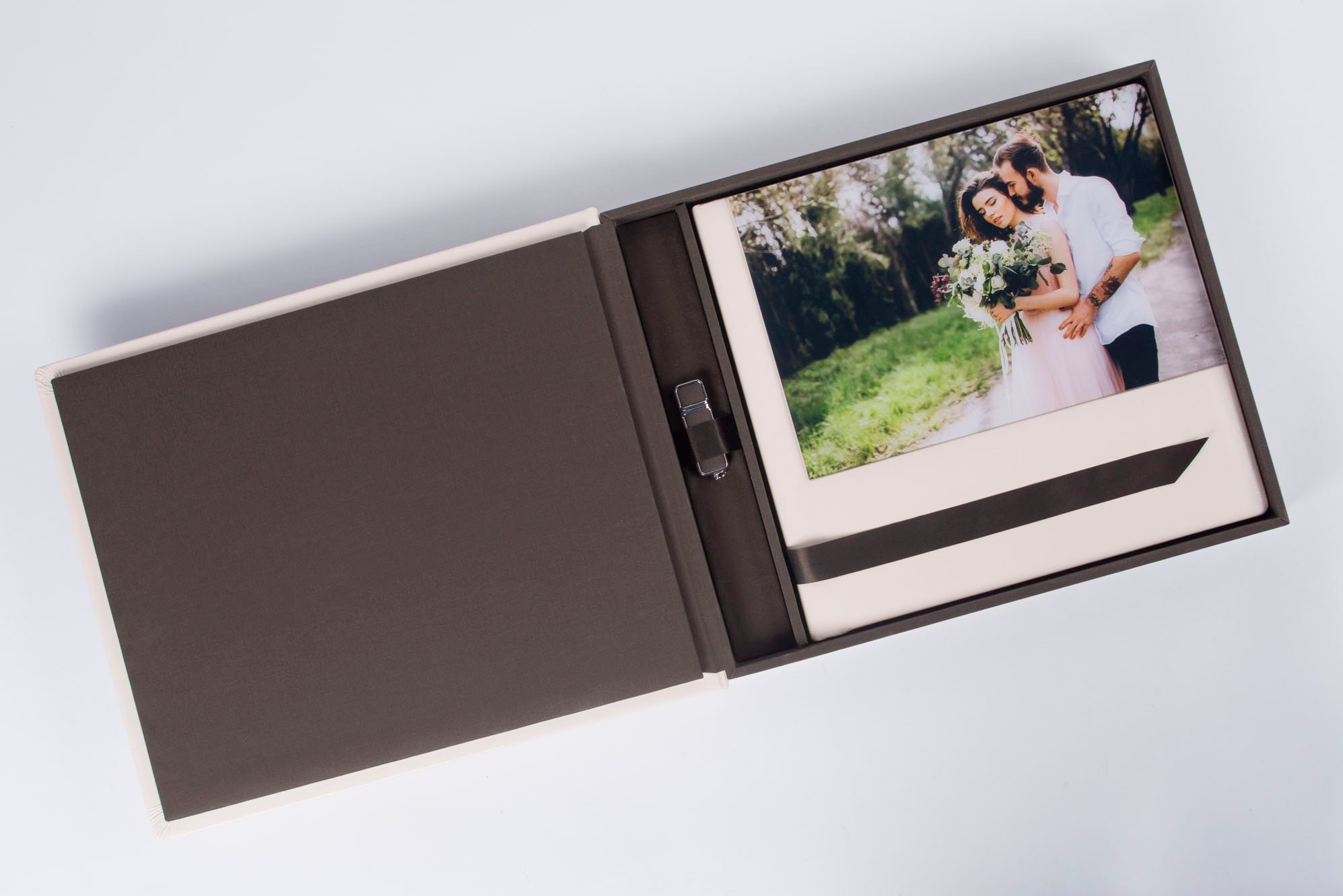 Otwarty, elegancki album fotograficzny z beżowo-brązowymi okładkami, zawierający zdjęcie pary młodej w plenerze oraz pendrive na czarnym pasku.