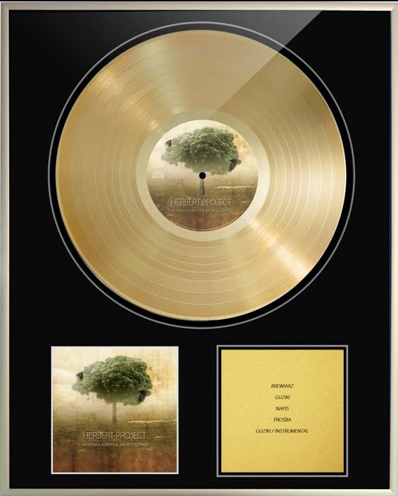 Złota płyta 'Herbert Project' w czarnej ramie, prezentowana w Płotach, z okładką albumu i listą utworów, sugerująca projekt graficzny i identyfikację wizualną.