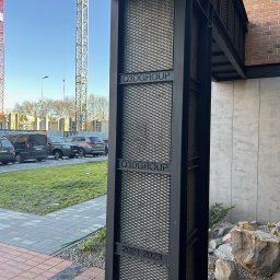 METAL-STYL Sp. z o.o. - Metalowa konstrukcja z siatką i napisem Q3DGROUP 2021-2023, stanowiąca element wejścia do budynku, z widocznymi elementami otoczenia, takimi jak chodnik, roślinność i samochody w tle.