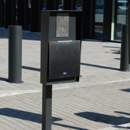 METAL-STYL Sp. z o.o. - Czarny, metalowy słupek z domofonem i skrzynką na listy na zewnątrz budynku na tle kostki brukowej i słupków zabezpieczających.