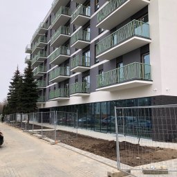 METAL-STYL Sp. z o.o. - Nowoczesny budynek mieszkalny z balkonami z zielonymi, metalowymi balustradami o ażurowym wzorze, widok z poziomu chodnika oddzielonego tymczasowym ogrodzeniem budowlanym.