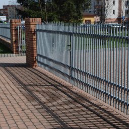 METAL-STYL Sp. z o.o. - Szara metalowa brama wjazdowa z pionowych prętów, osadzona pomiędzy ceglanymi słupkami na posesji z kostki brukowej.