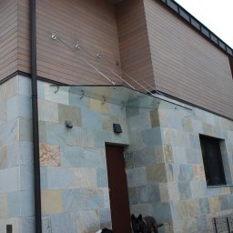 METAL-STYL Sp. z o.o. - Szklany daszek nad wejściem do domu z elewacją z kamienia naturalnego, podtrzymywany przez stalowe linki, widoczne dwa psy.